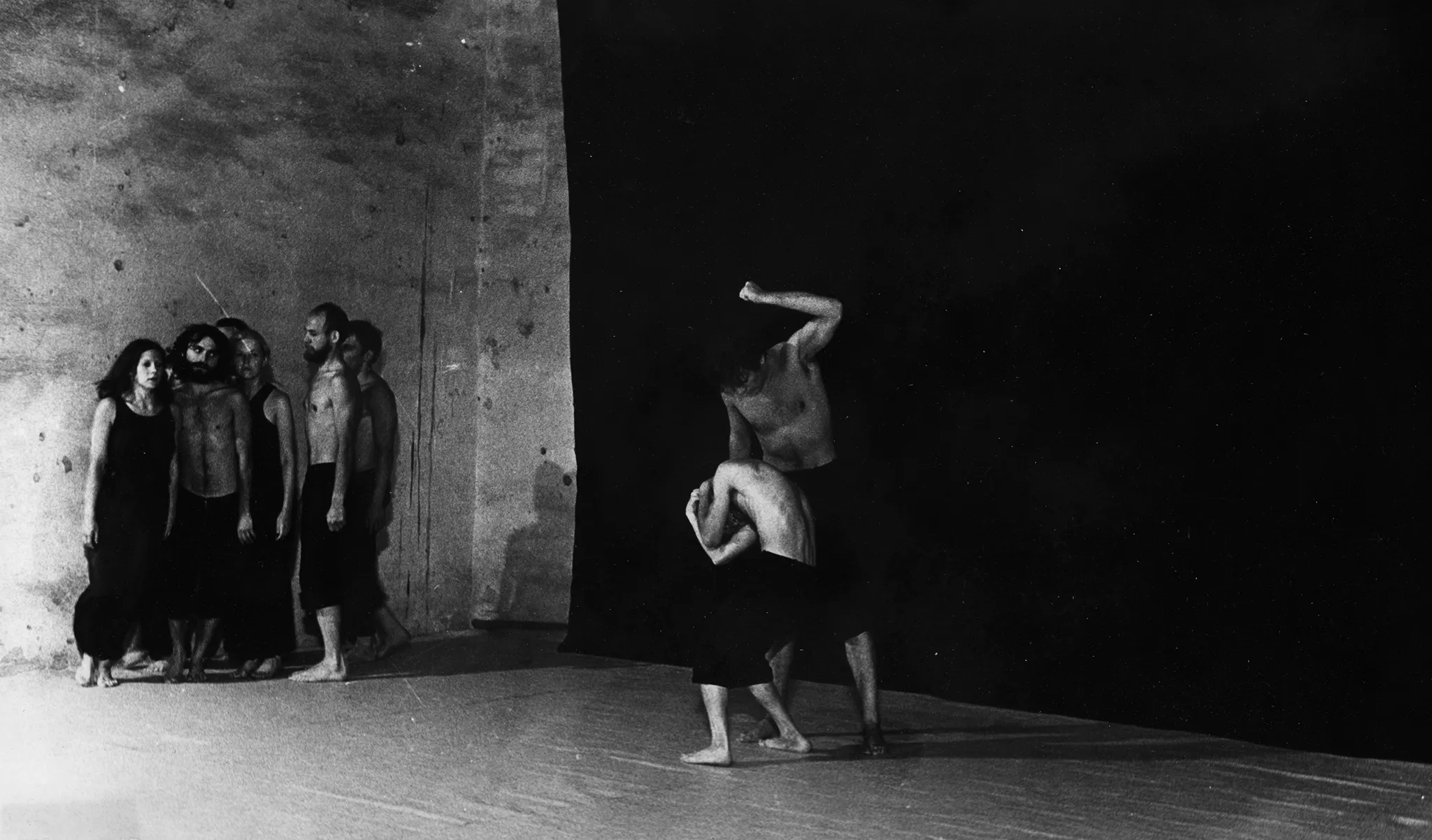 Po lewej stronie sceny na tle czarnej płachty stoi grupa aktorów i aktorek. Aktorzy ubrani są w czarne spodenki, mają nagie torsy. Aktorki ubrane w są w czarne spodnie i czarne koszulki. Przyglądają się dwóm aktorom w centrum. Jeden aktor wygląda, jakby bił drugiego. Bity aktor jest mocno skulony.