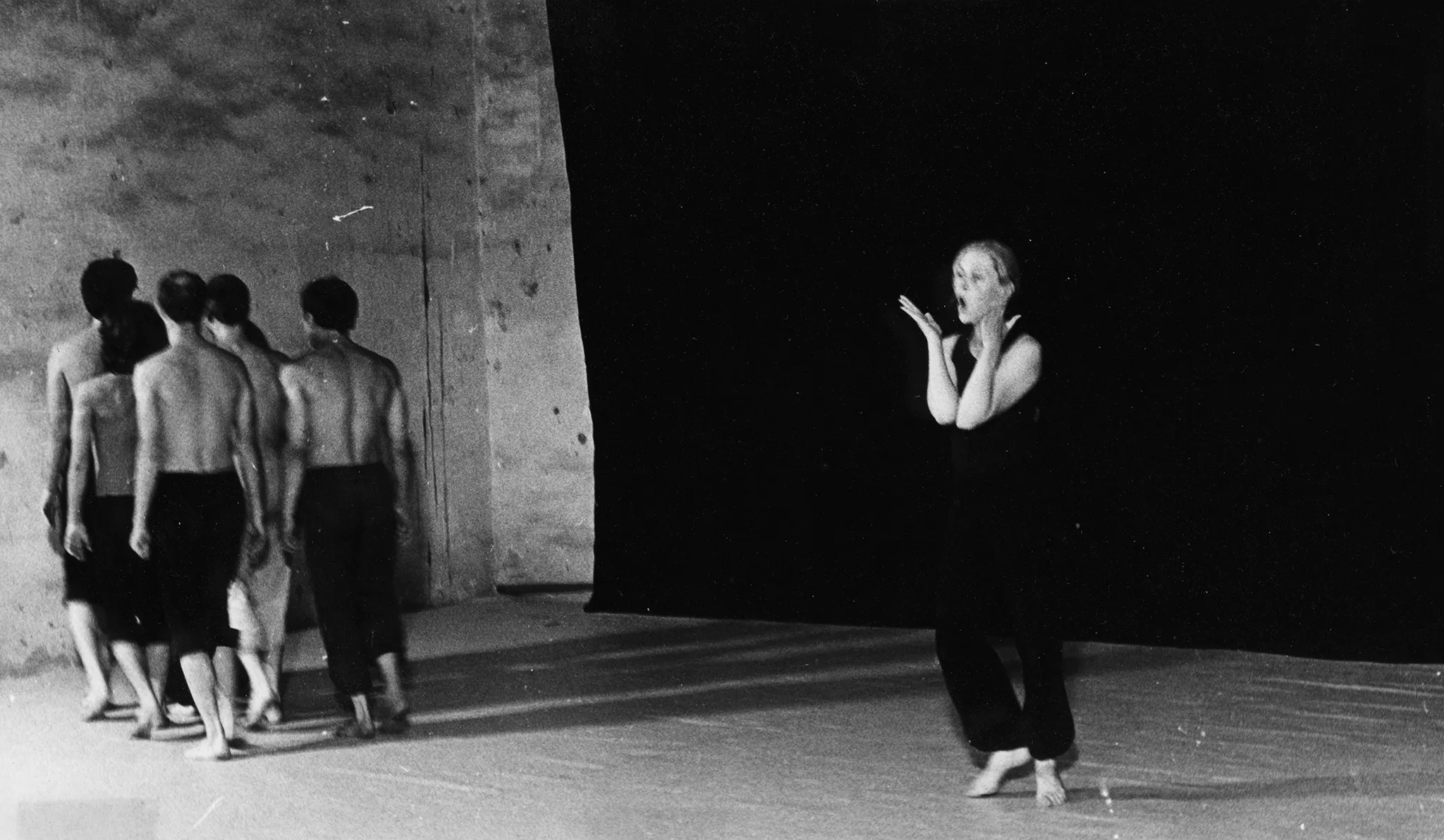 Po lewej stronie sceny na tle czarnej płachty stoi grupa aktorów odwróconych do widza plecami. Ubrani są w ciemne spodenki, mają nagie torsy. Są boso. W centrum stoi aktorka ubrana w czarne spodnie i czarną koszulkę na ramiączka. Jest boso. Wygląda, jakby biła się po twarzy.