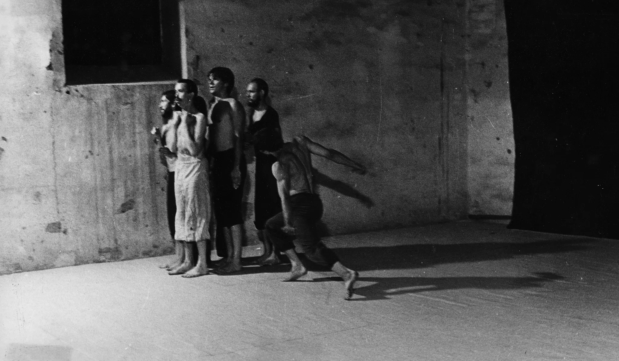 Po lewej stronie sceny na tle czarnej płachty z zwartej grupie stoją aktorzy. Na ugiętych nogach podchodzi do nich aktor. Wygląda, jakby chciał ich uderzyć.