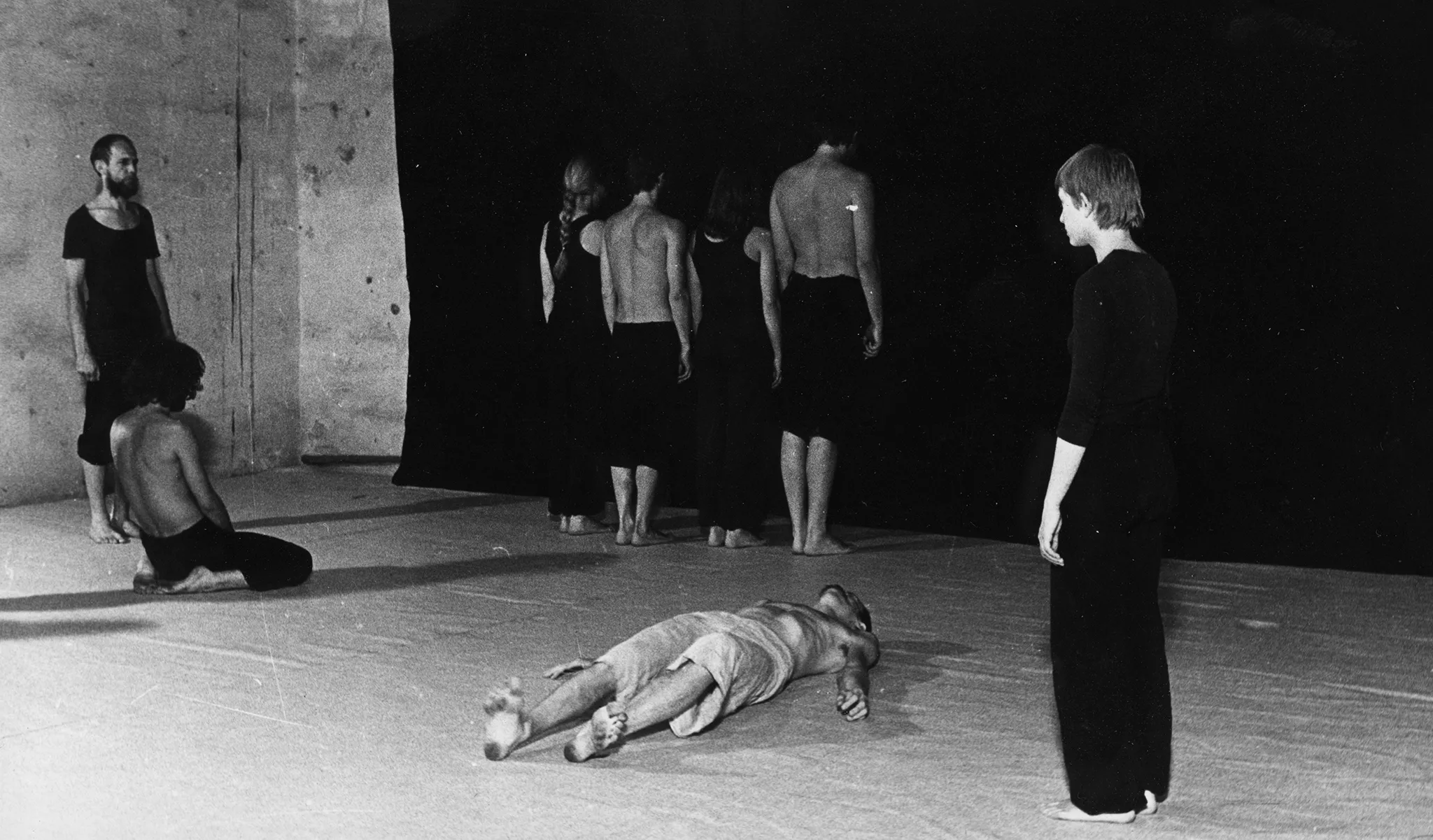 Grupa aktorów i aktorek na tle czarnej płachty. Po obu stronach naprzeciwko siebie stoją aktor i aktorka. Oboje są ubrani na czarno. Obok aktora klęczy aktor. Ubrany jest w czarne spodenki, jego tors jest nagi. Pod płachtą, plecami w stronę widza stoją aktorzy i aktorki. Aktorzy ubrani są w czarne spodenki, mają nagie torsy. Aktorki ubrane są w czarne spodnie i czarne koszulki na ramiączka. W centrum na plecach leży aktor. Ubrany jest w jasne spodenki, jego tors jest nagi.