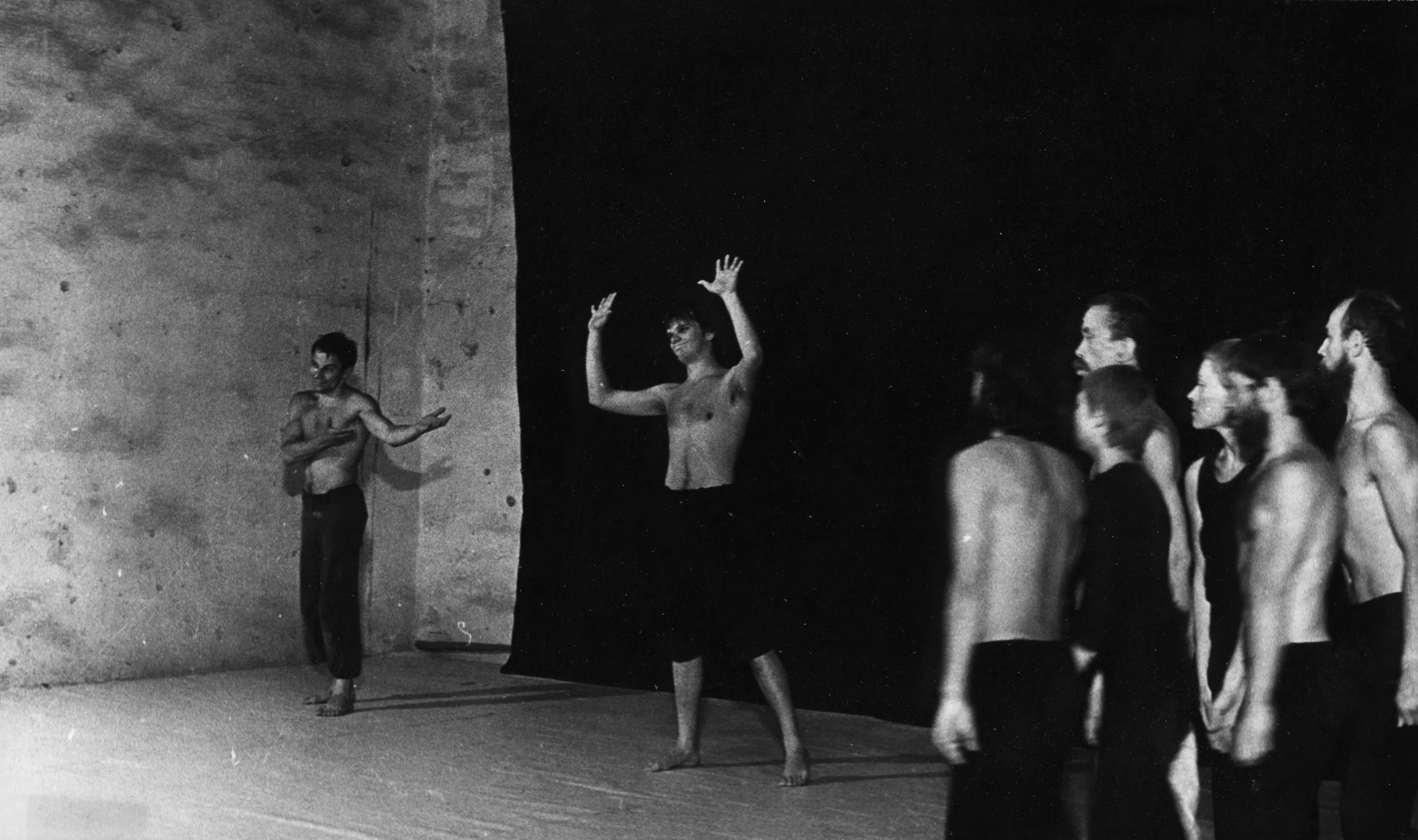 Po prawej stronie stoi grupa aktorów i aktorek zwróconych profilem w stronę widza. Aktorzy ubrani są w czarne spodenki, mają nagie torsy. Aktorki ubrane są w czarne spodnie i czarne koszulki. Po lewej stronie, pod czarną płachtą stoi dwóch aktorów. Obaj są ubrani w czarne spodnie i mają nagie torsy. Obaj się uśmiechają. Jeden aktor pokazuje wnętrza dłoni. Drugi aktor wskazuje na pierwszego aktora.