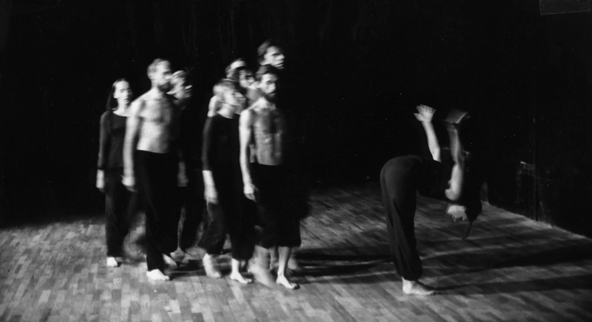 Rozmazane zdjęcie. Grupa aktorów i aktorek idzie przed siebie. Aktorzy są ubrani w czarne spodnie i mają nagie torsy. Aktorki ubrane są w czarne spodnie i czarne koszulki. Wszyscy są boso. Obok grupa stoi aktorka. Ma wyprostowane nogi i pochylony tułów. Aktorka trzyma ręce za plecami.