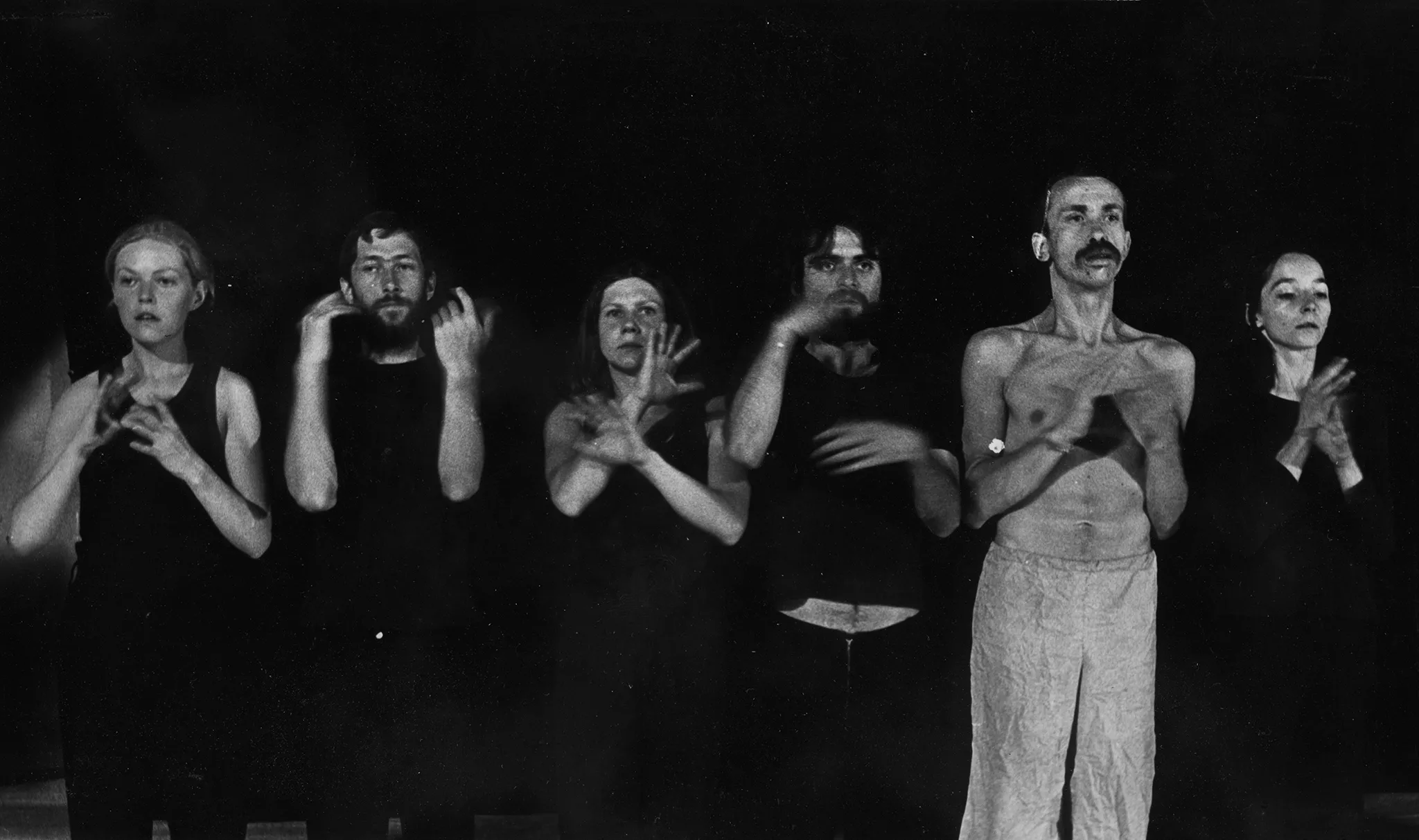 W szeregu, twarzą w stronę widza stoi grupa aktorów i aktorek. Wszyscy ubrani są na czarno poza aktorem, który jest ubrany w jasne spodenki i ma nagi tors. Wszyscy gestykulują dłońmi.