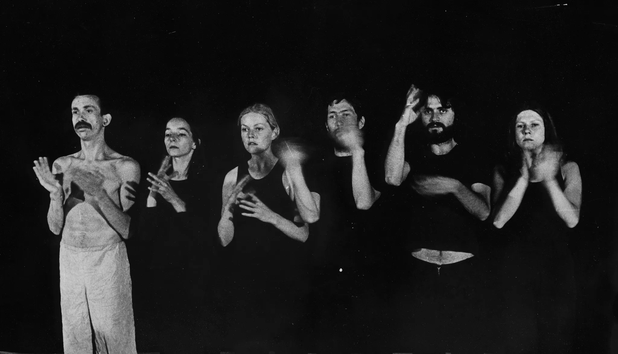 W szeregu, twarzą w stronę widza stoi grupa aktorów i aktorek. Wszyscy ubrani są na czarno poza aktorem, który jest ubrany w jasne spodenki i ma nagi tors. Wszyscy gestykulują dłońmi.