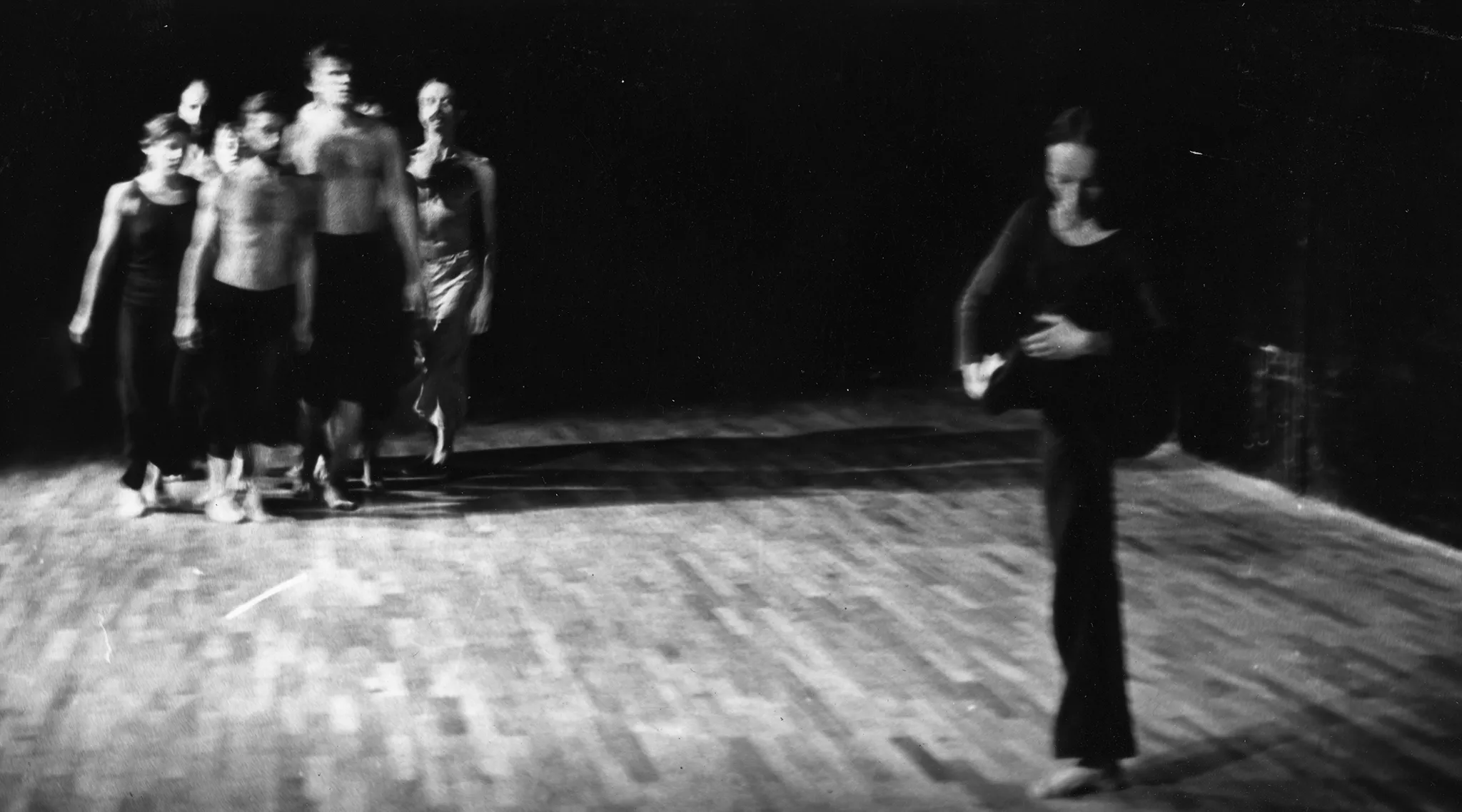 Rozmazane zdjęcie. Po lewej stronie idzie grupa aktorów i aktorek. Po prawej stronie na jednej nodze stoi aktorka. Jest ubrana na czarno.