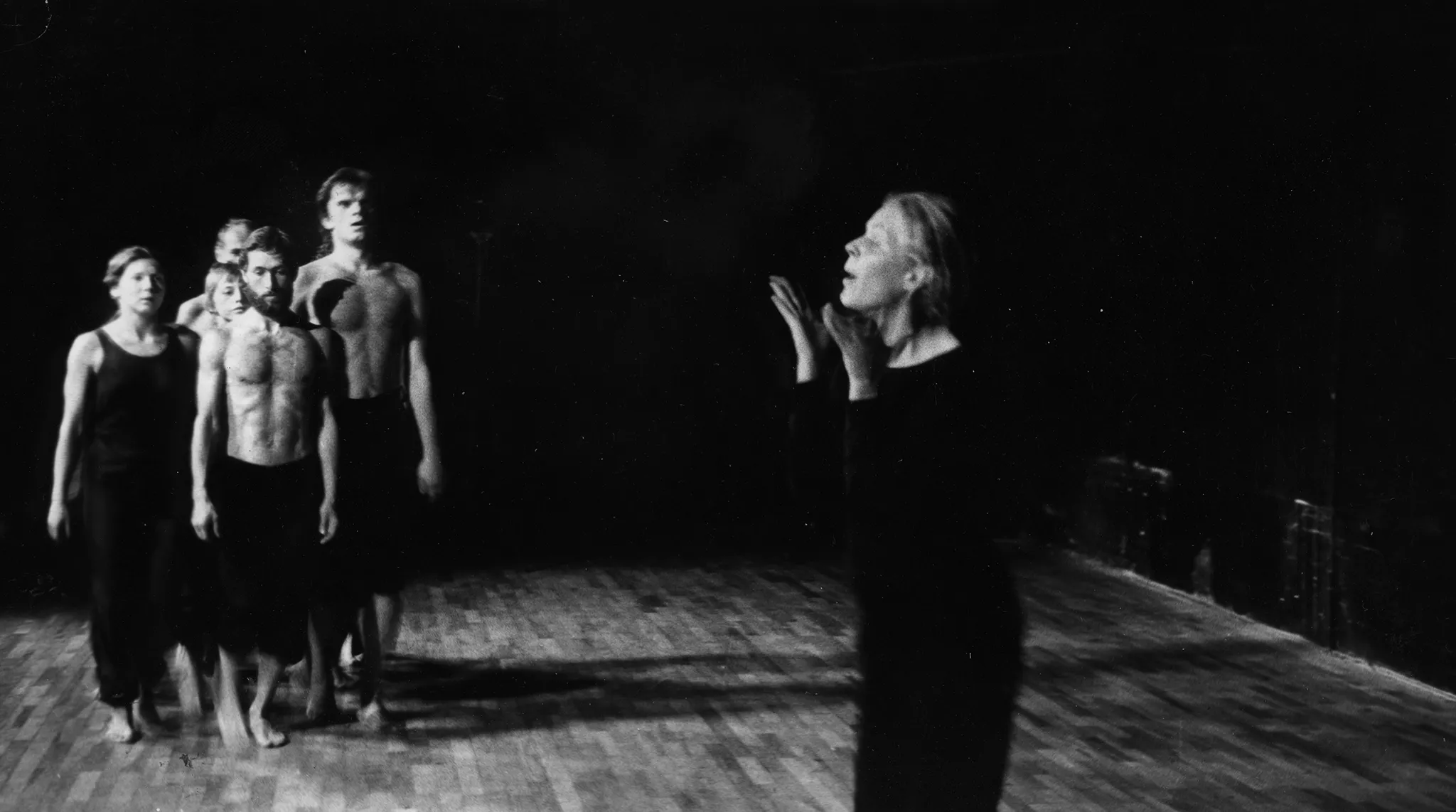 Po lewej stronie, zwrócona twarzą do widza, idzie grupa aktorów i aktorek. Aktorzy są ubrani w czarne spodnie i mają nagie torsy. Aktorki ubrane są w czarne spodnie i czarne koszulki. Po prawej stronie samotnie stoi widoczna od pasa w górę aktorka zwrócona do widza profilem. Aktorka dotyka dłońmi swojej twarzy. Ma otwarte usta.