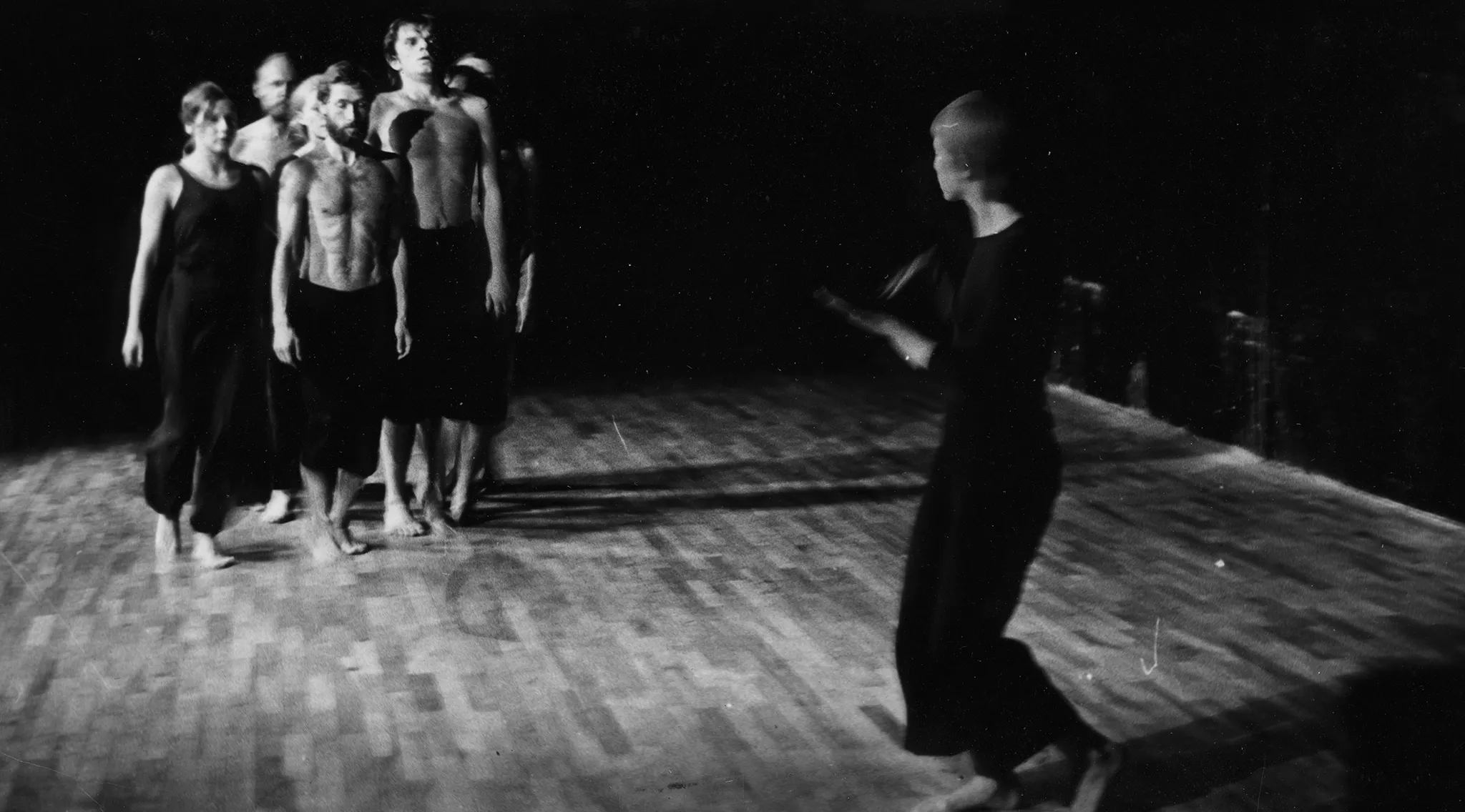 Po lewej stronie, zwrócona twarzą do widza, idzie grupa aktorów i aktorek. Aktorzy są ubrani w czarne spodnie i mają nagie torsy. Aktorki ubrane są w czarne spodnie i czarne koszulki. Po prawej stronie samotnie stoi aktorka. Jest ubrana na czarno, ma krótkie włosy. Wygląda, jakby tanczyła.
