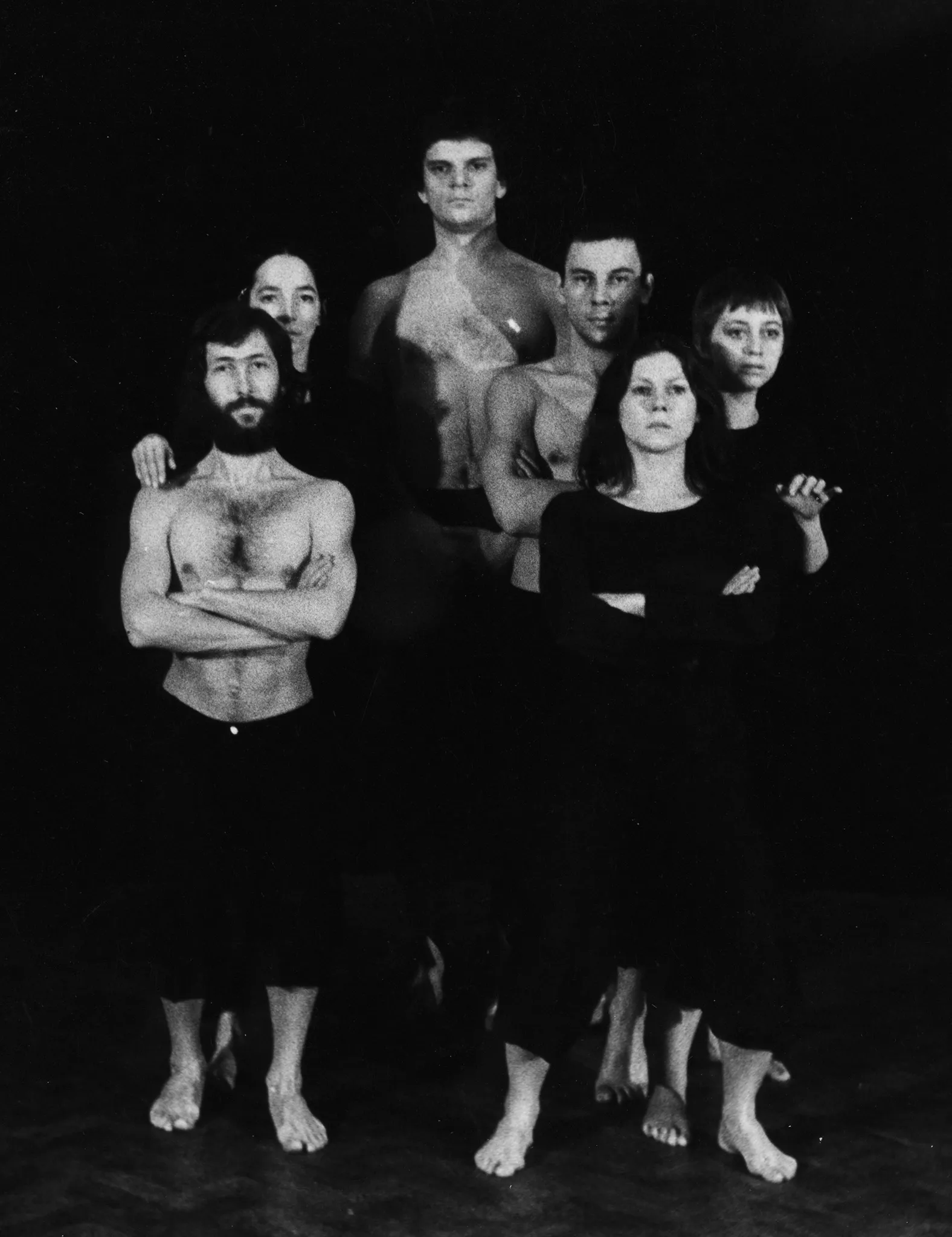 Grupa aktorów i aktorek stojących twarzą w stronę widza. Wyglądają, jakby pozowali do zdjęcia. Aktor i aktorka z przodu napinają mięśnie. Aktor jest ubrany w czarne spodenki i ma nagi tors. Aktorka ubrana jest w czarne sodnie i czarnę koszulkę. Za nimi stoją dwie aktorki i jeden aktor. Z tyłu stoi bardzo wysoki aktor.