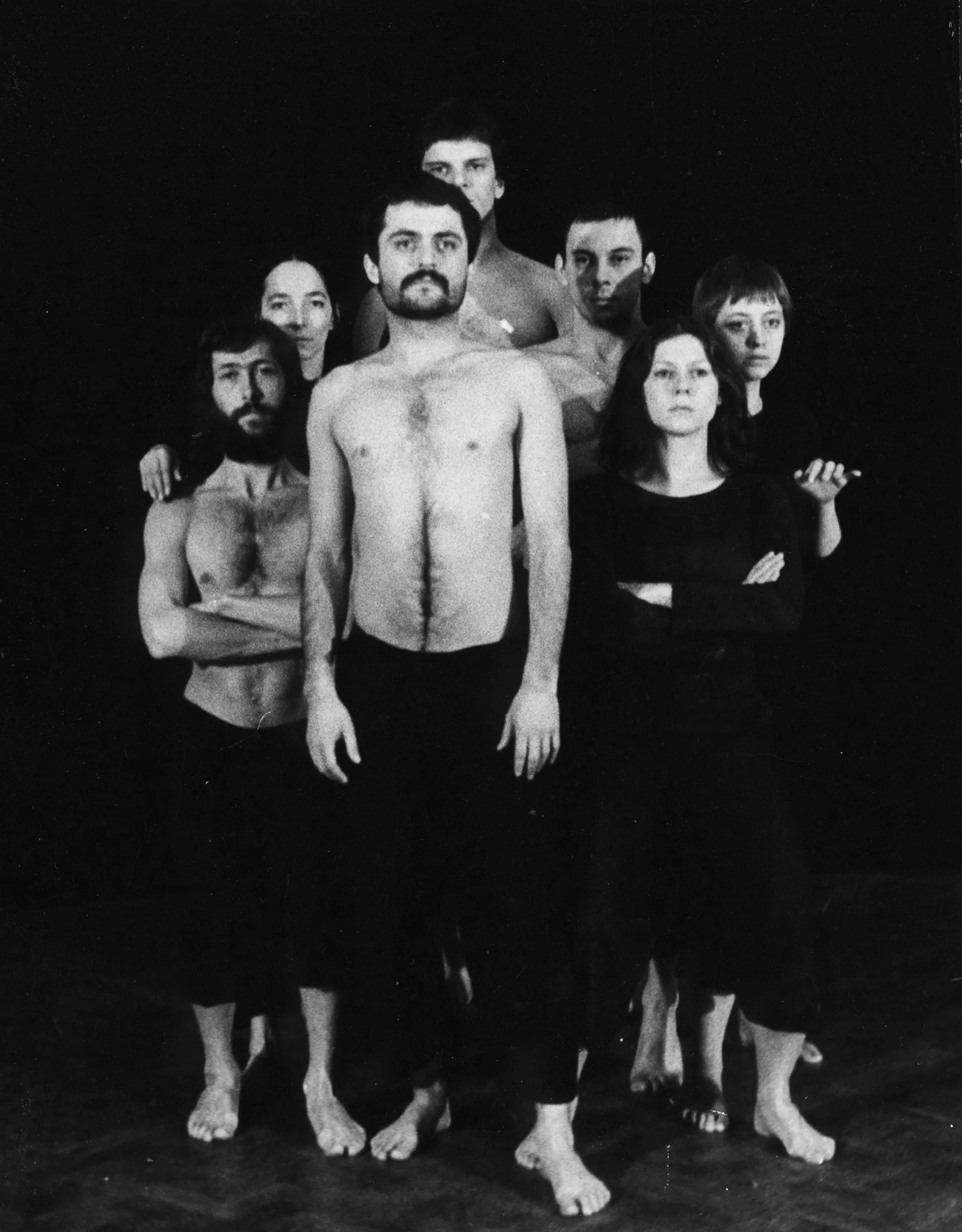 Grupa aktorów i aktorek stojących twarzą w stronę widza. Z przodu stoi aktor ubrany w czarne spodnie. Jego tors jest nagi. Za jego plecami stoi zwarta grupa aktorów i aktorek.