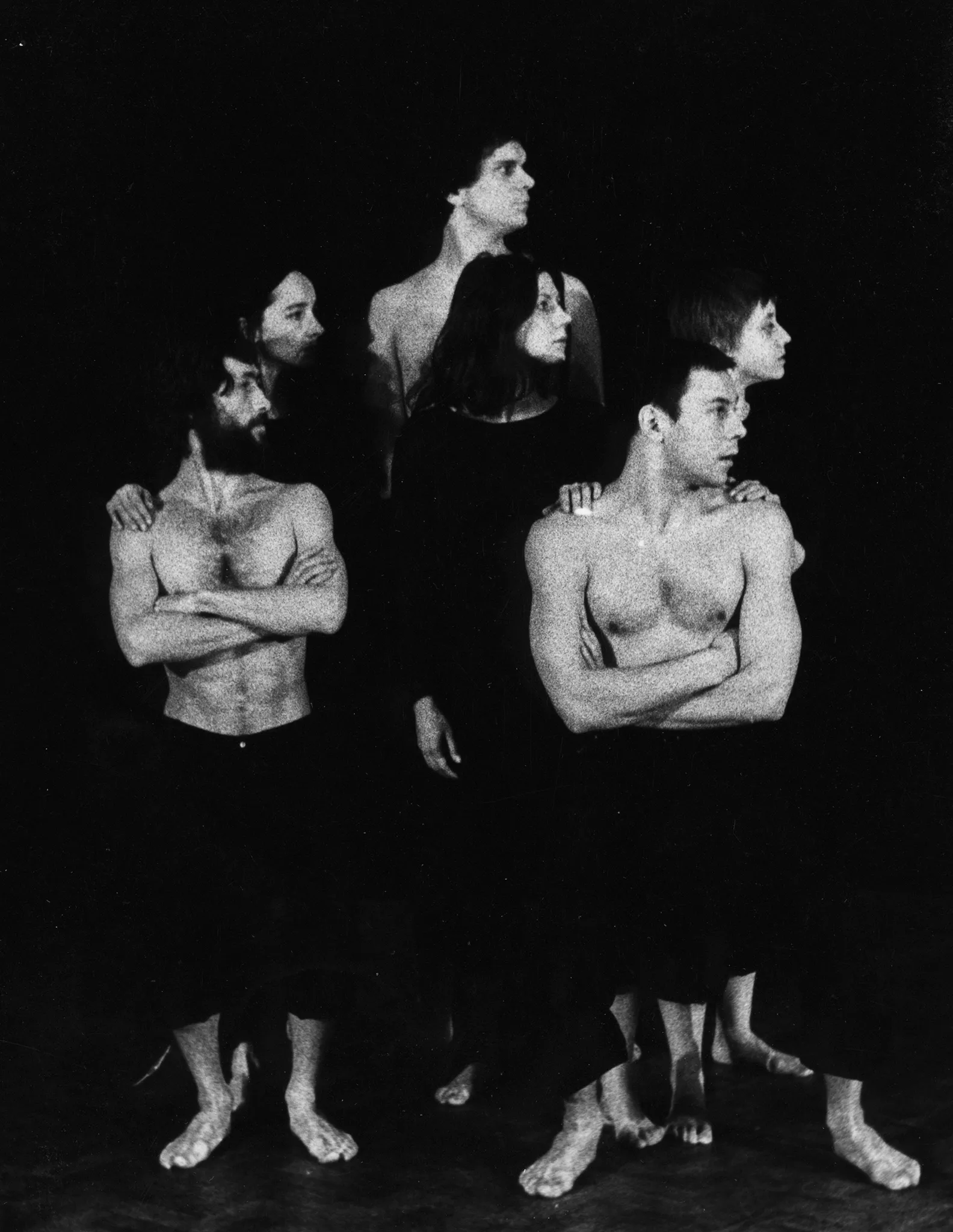 Grupa aktorów i aktorek stojących profilem w stronę widza. Wyglądają, jakby pozowali do zdjęcia. Dwóch aktorów z przodu napina mięśnie. Ubrani są w czarne spodenki i mają nagie torsy. Za nimi stoją trzy aktorki ubrane na czarno. Z tyłu stoi bardzo wysoki aktor.