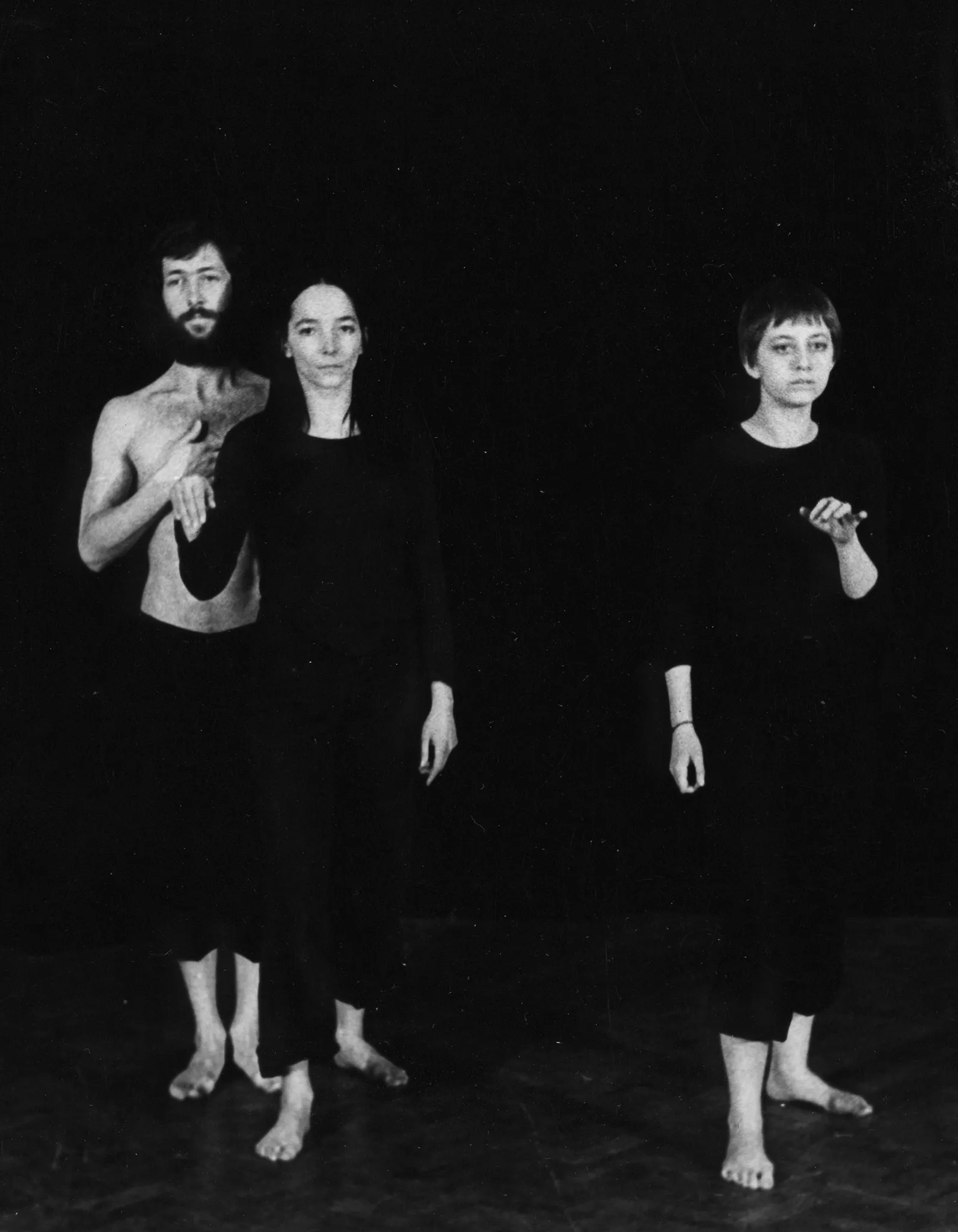 Jeden aktor i dwie aktorki. Stoją zwróceni twarzą w stronę widza, jakby pozowali do zdjęcia. Po lewej stronie aktor i aktorka stoją bardzo blisko siebie. Aktorka po prawej stronie stoi samotnie. Aktor ubrany jest w czarne spodenki i ma nagi tors. Aktorki ubrane są w czarne spodnie i czarne koszulki. Wszyscy są boso.