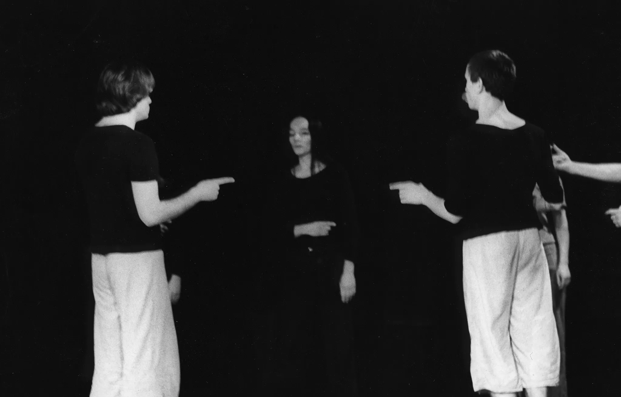 Grupa aktorów i aktorek stoi w okręgu. Wskazują na siebie palcami. Dwóch aktorów na pierwszym planie jest odwróconych plecami w stronę widza. Ubrani są w jasne spodnie i czarne koszulki. Na drugim planie stoi aktorka ubrana na czarno.