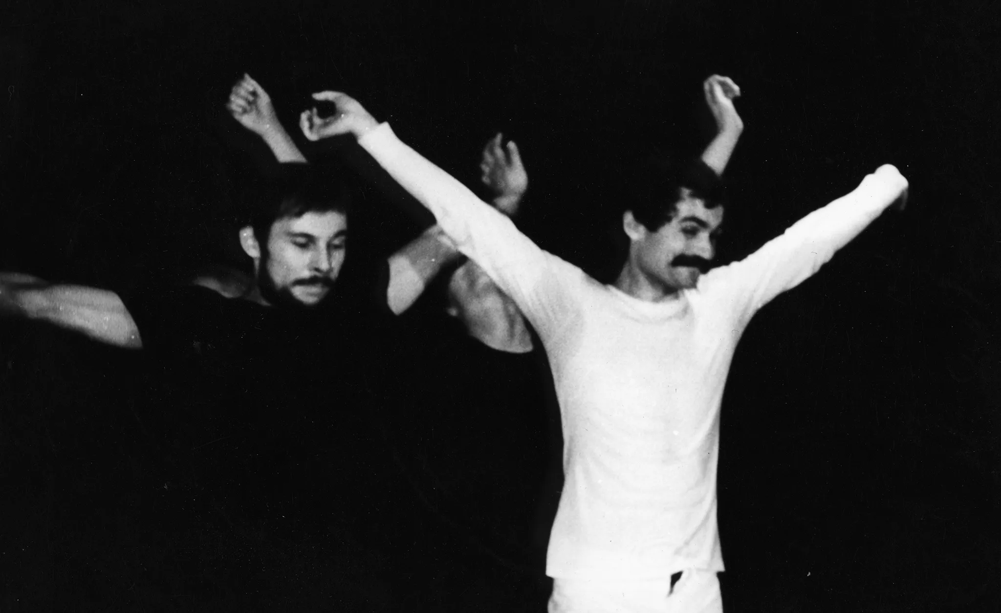 Grupa aktorów stoi blisko siebie. Mają ręce szeroko wyciągnięte na boki. Na pierwszym planie widać dwóch aktorów. Jeden ubrany jest na czarno, drugi na biało. Aktor ubrany na czarno jest poważny, aktor ubrany na biało się uśmiecha.