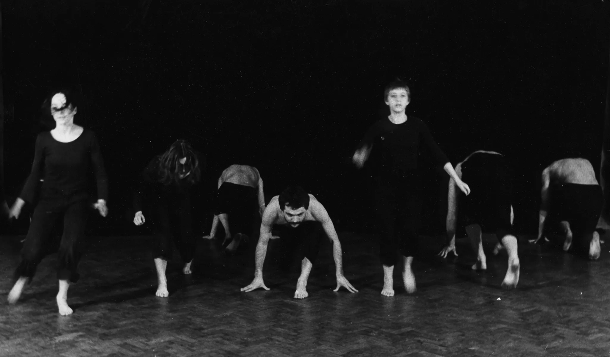 Grupa aktorów i aktorek wygląda, jakby przygotowywała się do biegu. Jeden aktor zwrócony jest twarzą w stronę widza, reszta zwrócona jest do widza plecami. Wyróżniają się trzy aktorki, które stoją. Są zwrócone twarzą w stronę widza. Ubrane są na czarno.