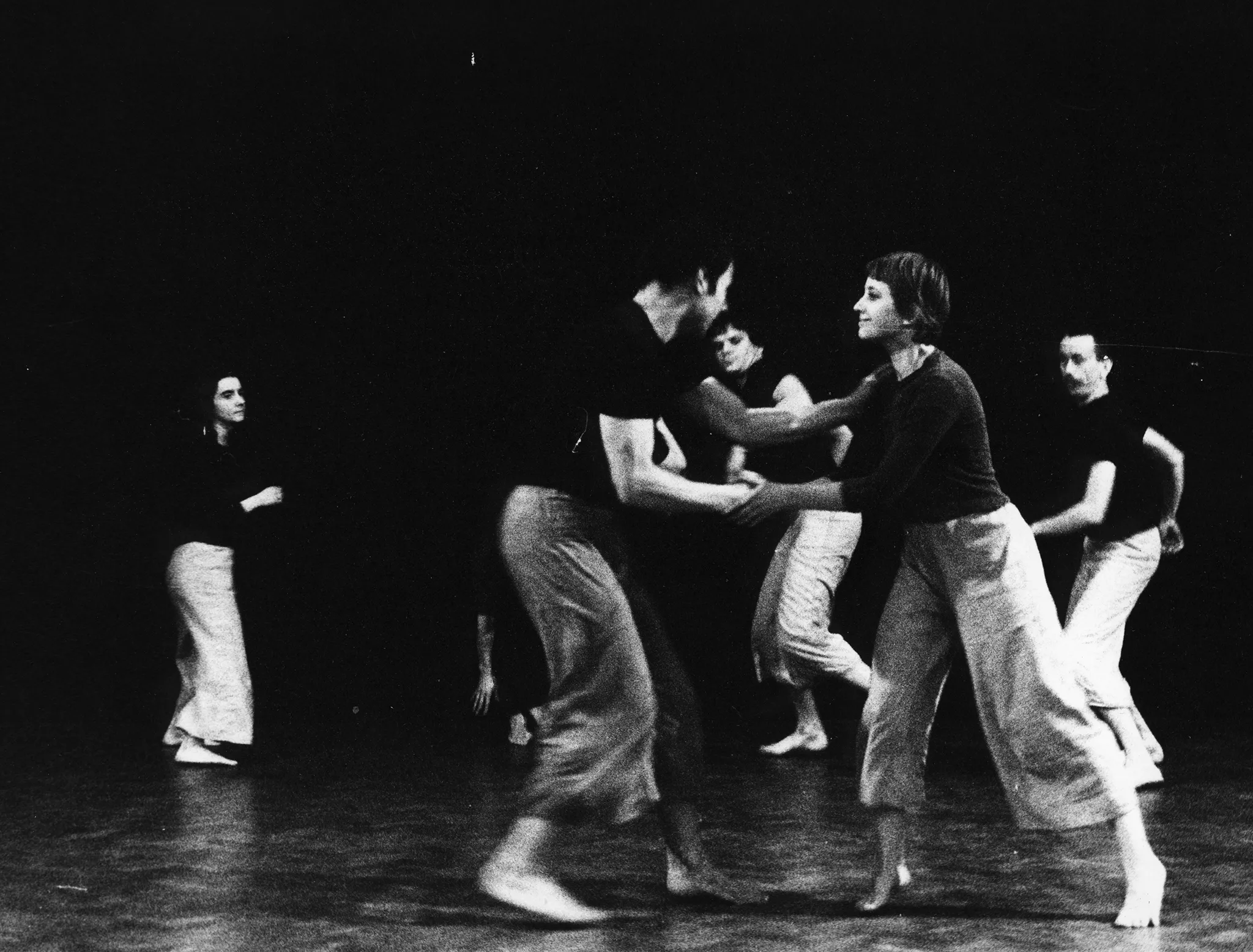 Grupa aktorów i aktorek energicznie porusza się na scenie. Wszyscy ubrani są w jasne spodnie i czarne koszulki. Na pierwszym planie w bliskim kontakcie stoją aktor i aktorka. Uśmiechają się do siebie i wyglądają, jakby tańczyli.