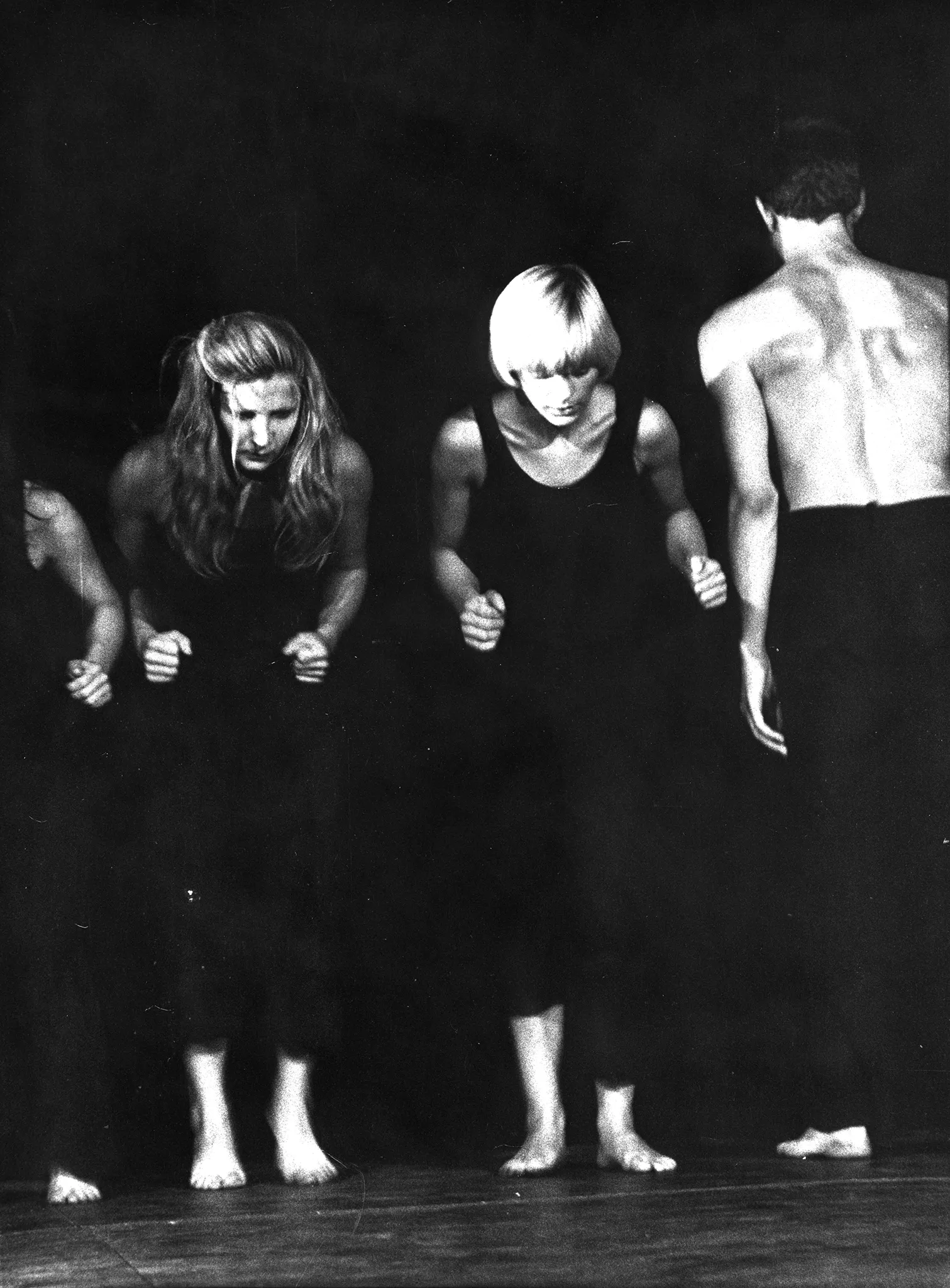 Dwie aktorki stoją zwrócone twarzami w stronę widza. Są lekko pochylone. Ubrane są w czarne spodnie i czarne koszulki na ramiączka. Są boso. Obok nich stoi zwrócony plecami w stronę widza aktor. Ubrany jest w czarne spodnie i ma nagi tors. Jest boso.