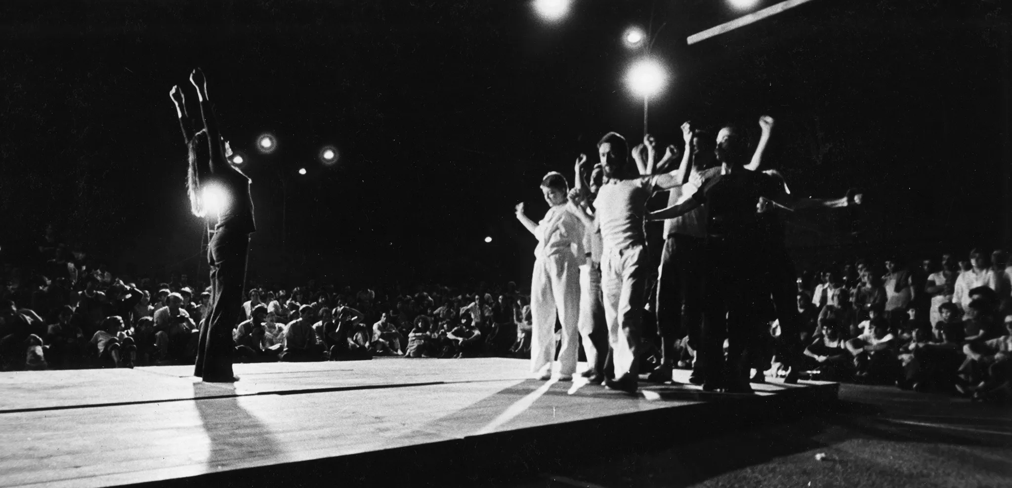 Scena plenerowa. Na krańcu sceny stoi grupa aktorów i aktorek. Uginają ręce, jakby napinali mięśnie. Naprzeciwko grupe stoi aktorka z wysoko wyciągniętymi rękami. Grupie aktorów przygląda się duża grupa widzów. Działania aktorów oświetalają latarnie.