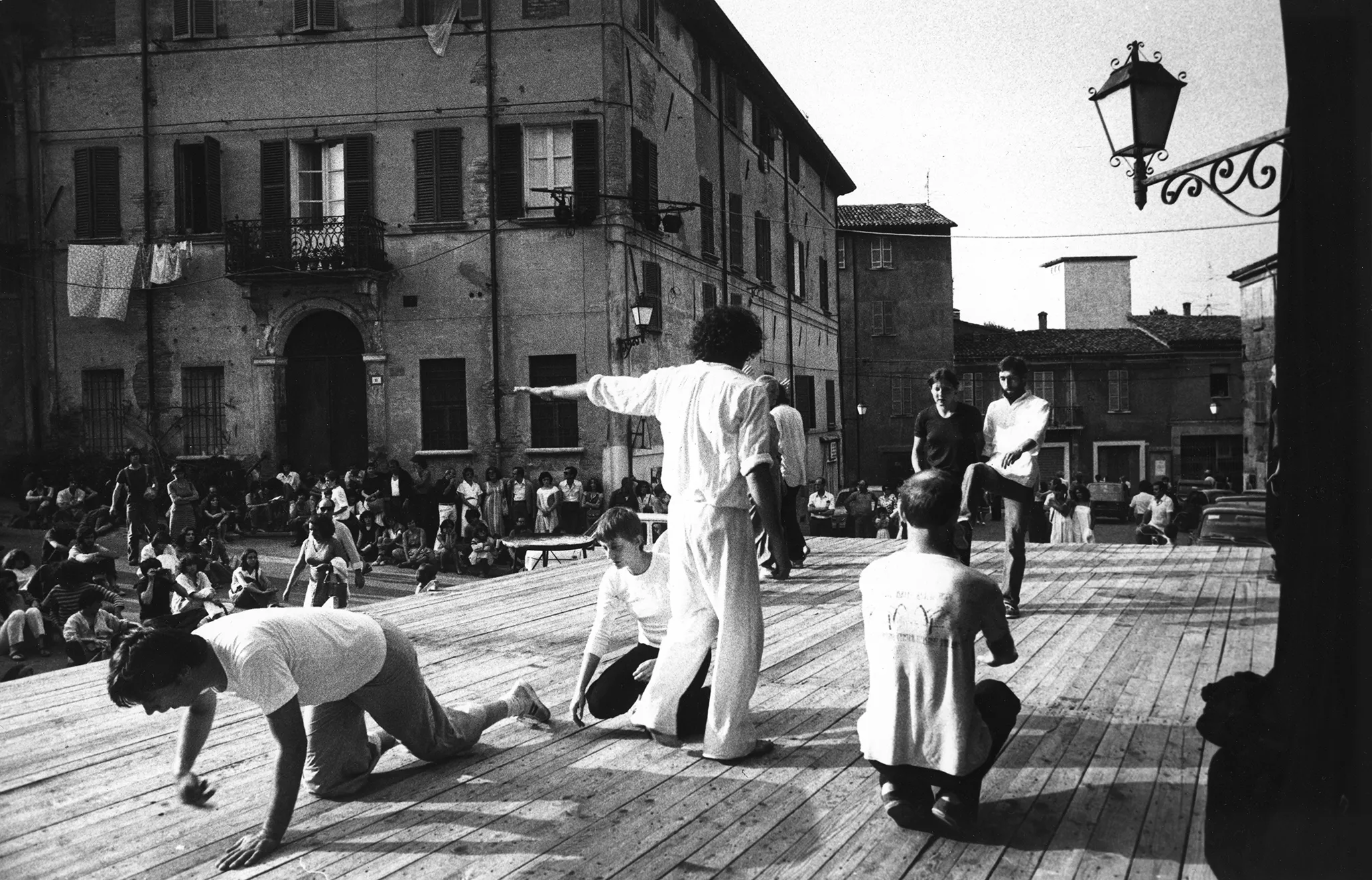 Scena ustawiona w przestrzeni miejskiej. Na scenie grupa aktorów i aktorek ubranych na biało. Wyróżnia się jedna aktorka ubrana na czarno. Aktorzy i aktorki wykonują indywidualne działania ruchowe. W tle widać budynki mieszkalne. Pod sceną oraz pod budynkami siedzą widzowie.