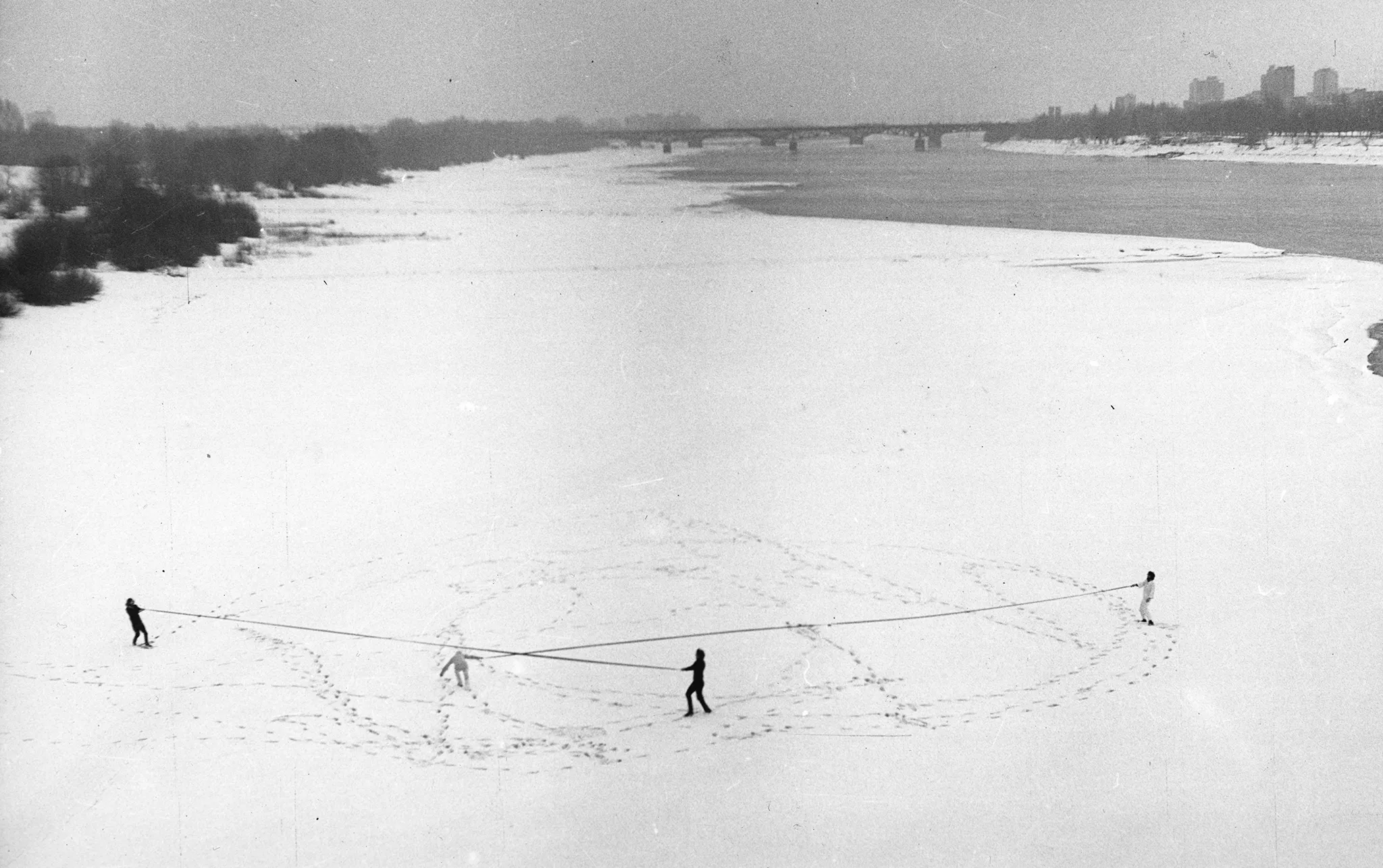 Zaśnieżona plaża przy rzece. W oddali widać most i miasto. Na plaży czworo aktorów w zimowych ubraniach rozciąga dwie długie liny. Na śniegu widać ślady ich butów.