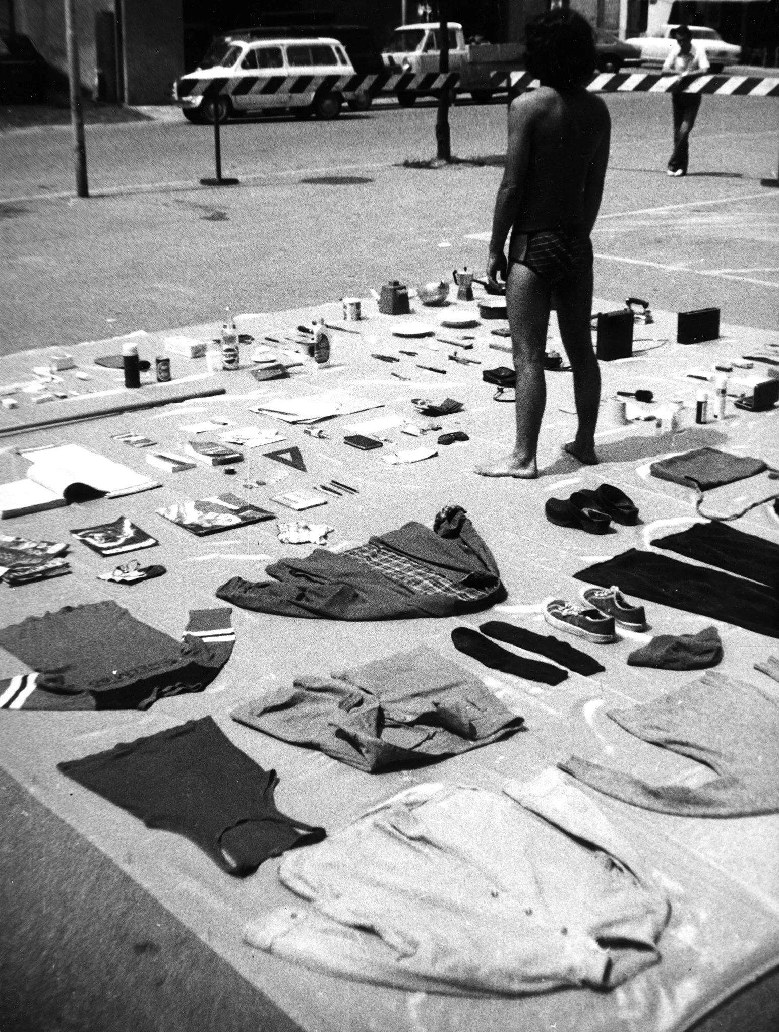 Przestrzeń miejska. Przy ulicy leży rozłożona płachta, na której leżą przedmioty, między innymi kurtki, koszulki, buty, butelki, przybory szkolne. Na płachcie stoi odwrocony plecami do widza aktor. Ubrany jest tylko w majtki. W oddali widać samochody.