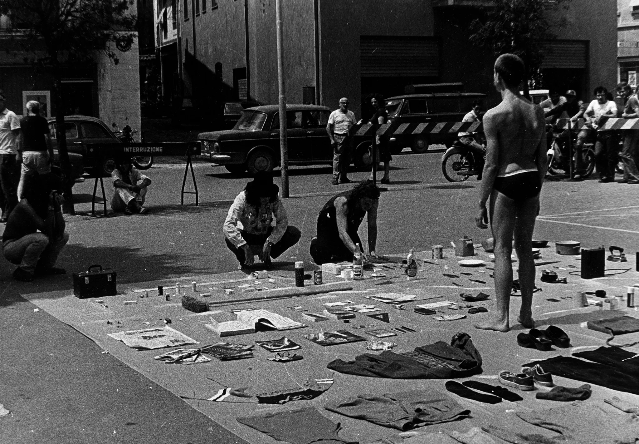 Przestrzeń miejska. Przy ulicy leży rozłożona płachta, na której leżą przedmioty, między innymi kurtki, koszulki, buty, butelki, przybory szkolne. Na płachcie stoi odwrocony plecami do widza aktor. Ubrany jest tylko w majtki. Przy płachcie i przy ulicy stoją widzowie.