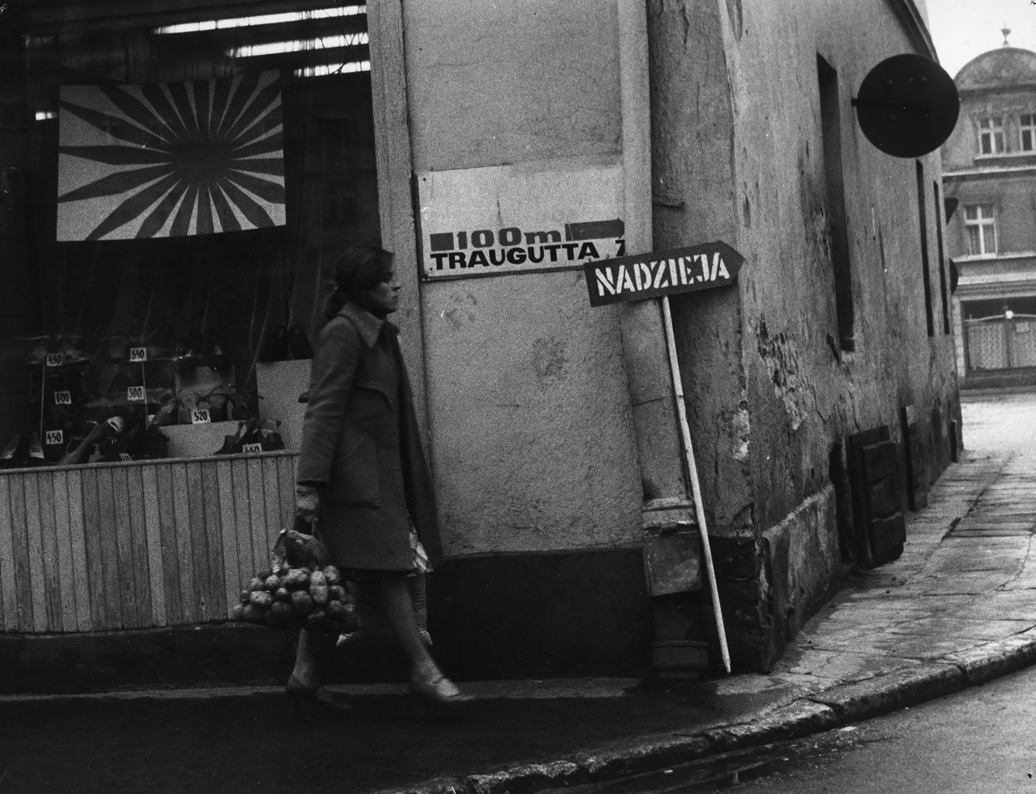 Przestrzeń miejska. Róg budynku z witryną wystawową. Na budynku widnieje napis "100 m Traugutta" i strzałka. O budynek stoi oparta strzałka z napisem "NADZIEJA". Pod budynkiem przechodzi kobieta z siatką pełną ziemniaków. Ubrana jest w płaszcz.