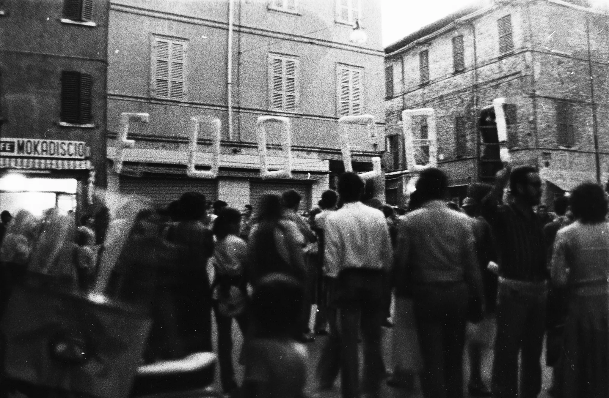 Niewyraźne zdjęcie. Przestrzeń miejska. Wśród budynków stoi tłum ludzi zwróconych w stronę widza plecami. Nad głowami ludzi wisi napis "FUOCO!"