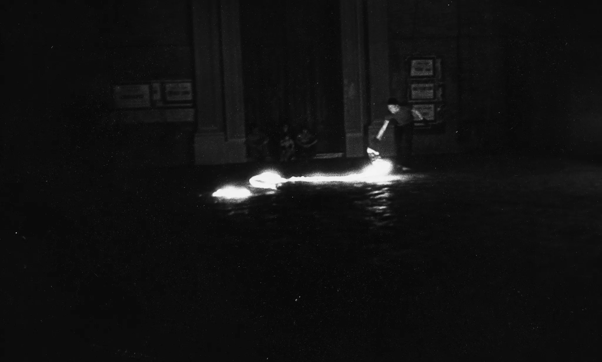 Niewyraźne zdjęcie. Przestrzeń miejska. Na ulicy stoi aktor. Wygląda, jakby coś podpalał. Za jego plecami widać budynek, pod którym siedzi troje widzów.