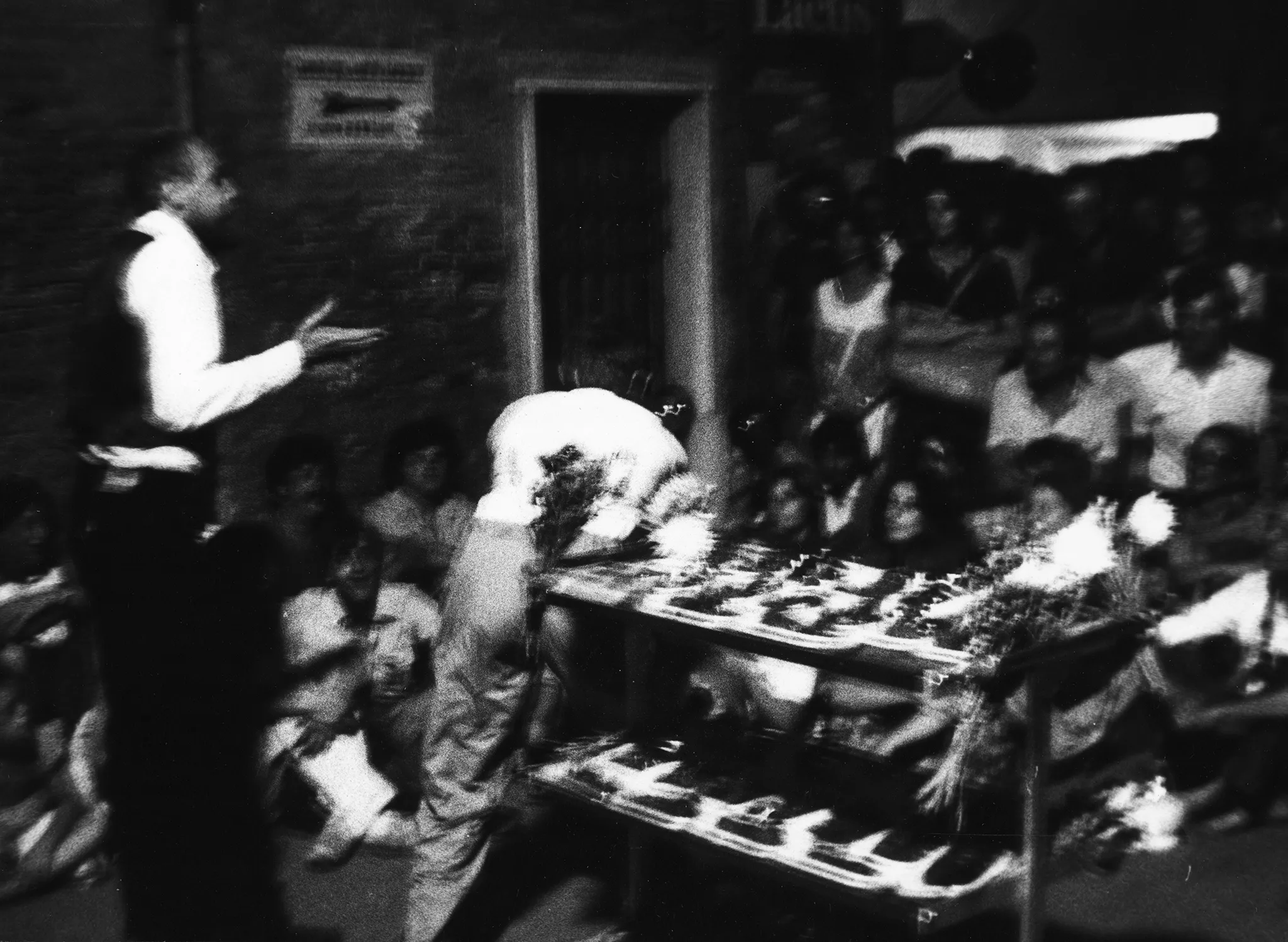 Niewyraźne zdjęcie. Aktor zwrócony plecami do widza przemawia do publiczności. Obok niego stoi kwietnik.