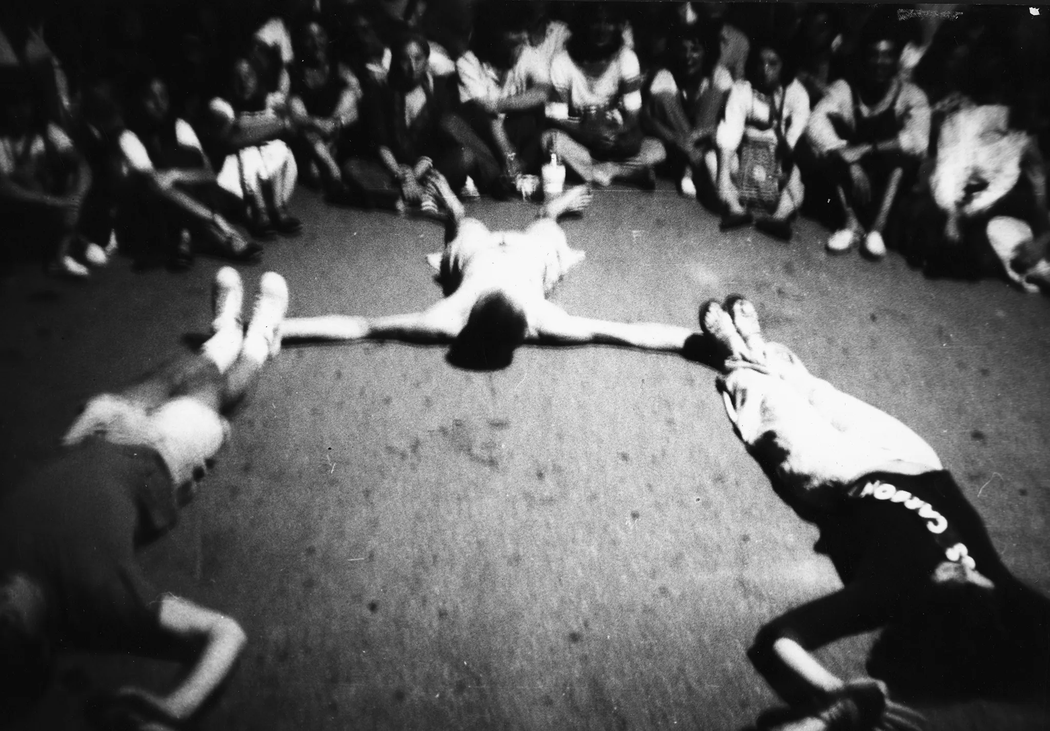 Niewyraźne zdjęcie. Trzech aktorów leży krzyżem plecami na połodze. Na drugim planie siedzi grupa widzów przyglądających się aktorom.