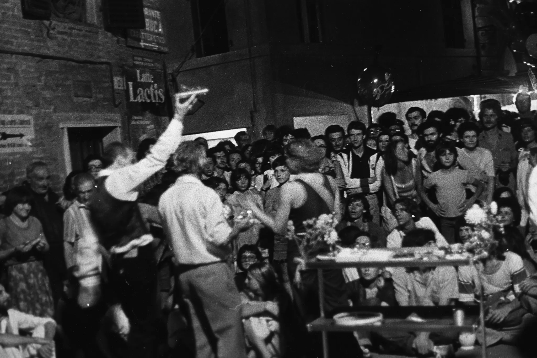 Przestrzeń miejska. Dwoje aktorów i jedno aktorka zwróconych plecami w stronę widza przemawia do publiczności. Obok nich stoi kwietnik.