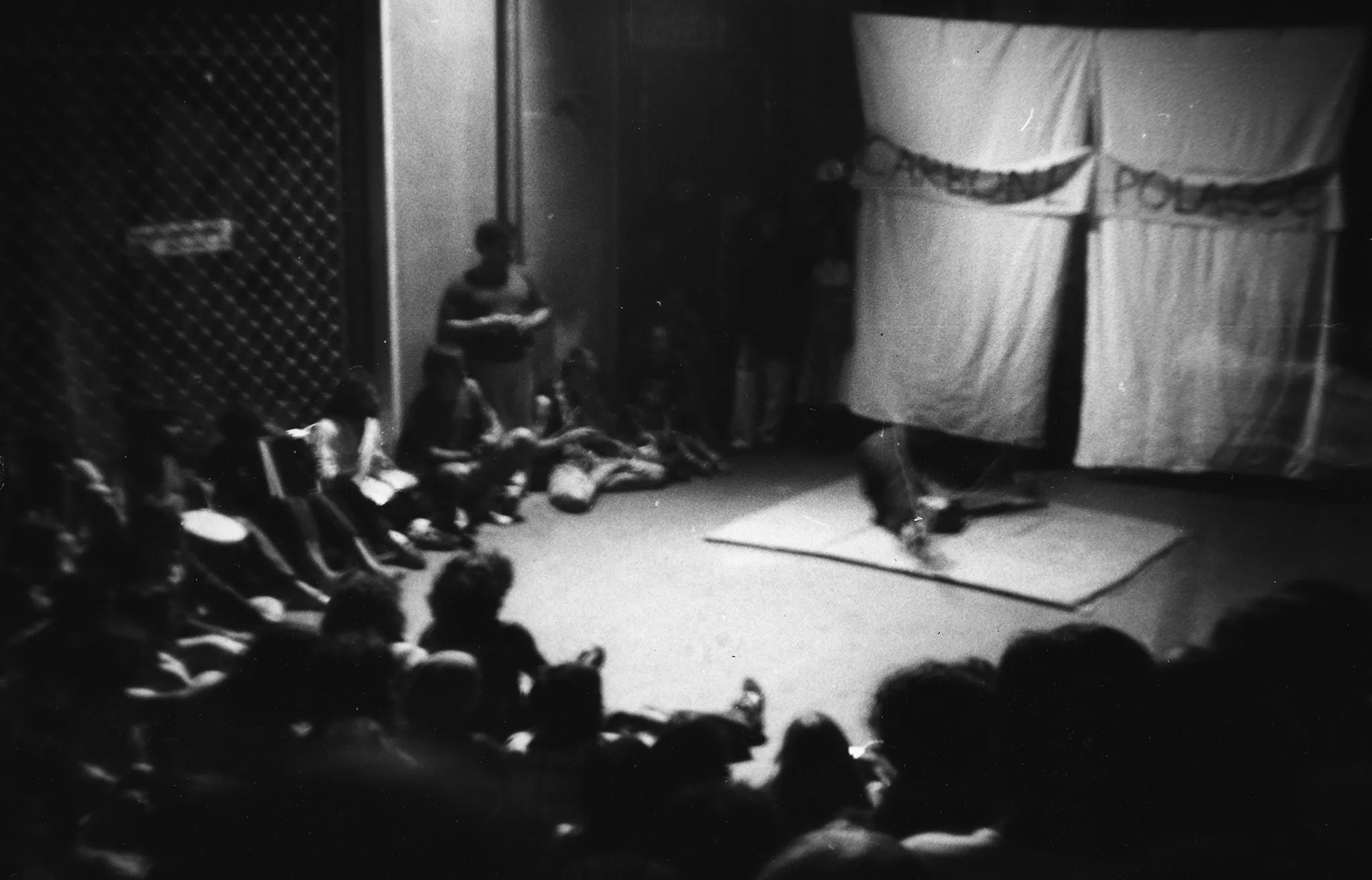 Niewyraźne zdjęcie. Grupa widzów siedzi zgromadzona przy scenie. Na scenie leży jasna mata. Z tyłu sceny wisi biała kurtyna. Na kurtynie wisi biała szarfa z czarnym napisem "CARBONE POLACCO".