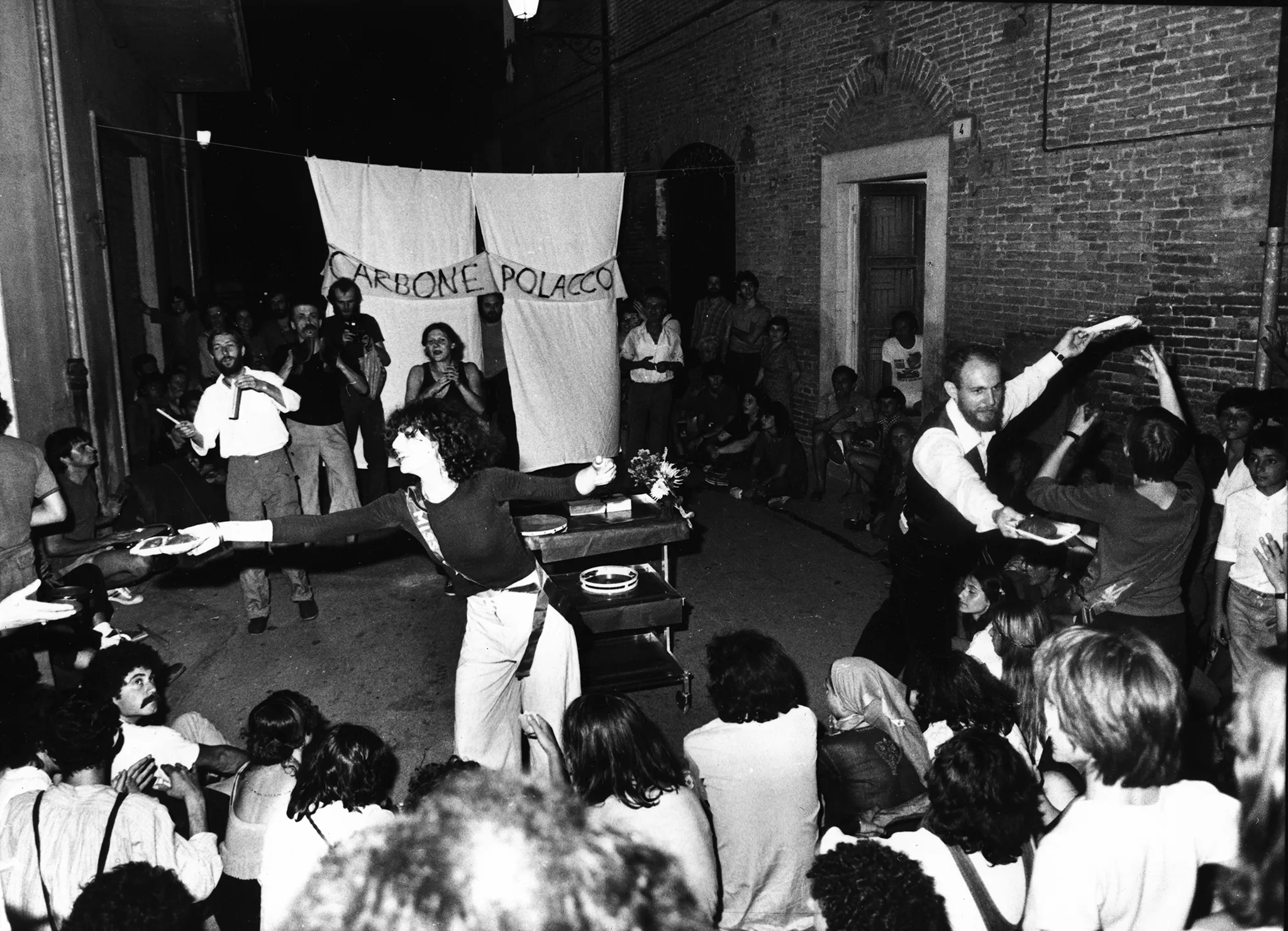 Przestrzeń miejska. Między budynkami siedzi grupa widzów zwróconych do nas plecami. Przed nimi stoi aktorzy i aktorki rozdający im przedmioty na tackach. Z tyłu wisi biała kurtyna. Na kurtynie wisi biała szarfa z czarnym napisem "CARBONE POLACCO".