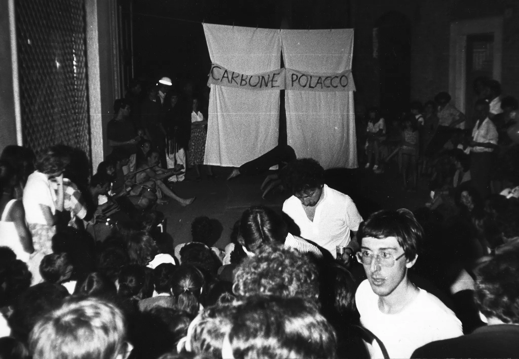 Przestrzeń miejska. Między budynkami zgromadzona publiczność. Z tyłu wisi biała kurtyna. Na kurtynie wisi biała szarfa z napisem "CARBONE POLACCO". Pod kurtyną aktorka wykonuje akrobatyczną pozę.