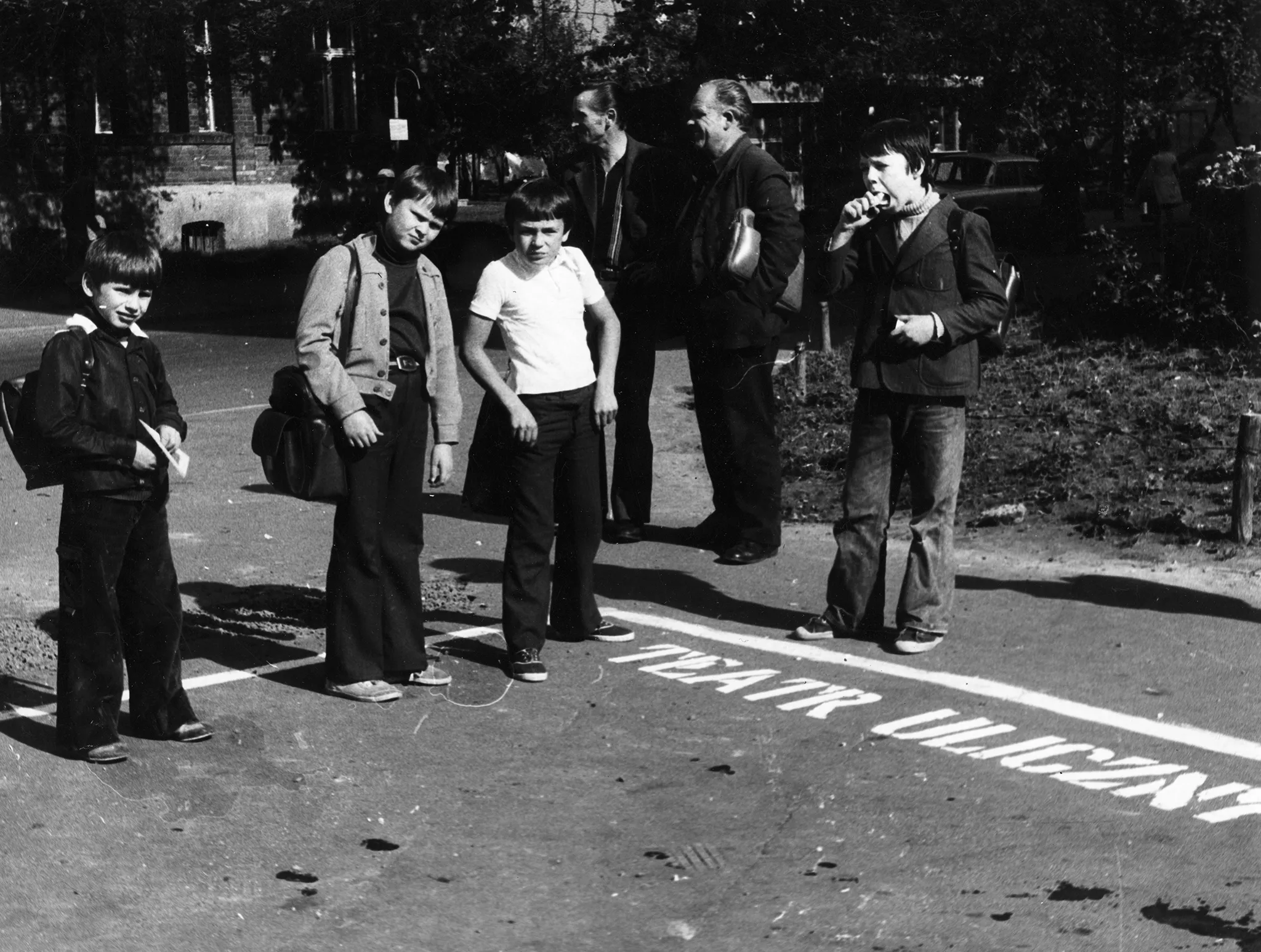 Przestrzeń miejska. Czterech chłopców wygląda, jakby pozowało do zdjęcia. Mają skrzywione miny. Za nimi stoi dwóch mężczyzn zwróconych profilem w stronę widza. Na chodniku widać biały napis "TEATR ULICZNY".