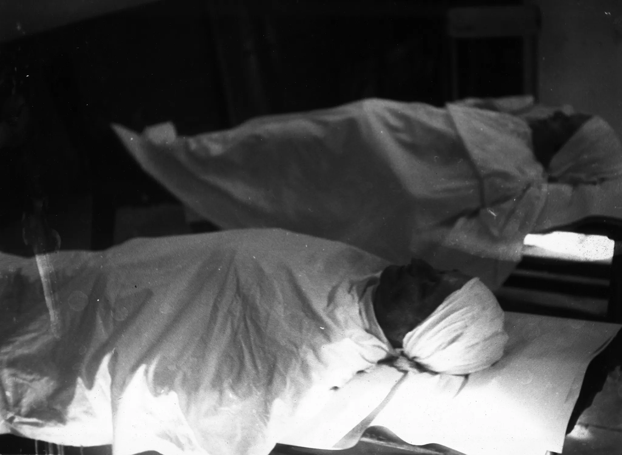 Obok siebie stoją dwa łóżka wyglądające na szpitalne. Na każdym łóżku leży osoba z owiniętą bandażem głową i przykryta białą płachtą.