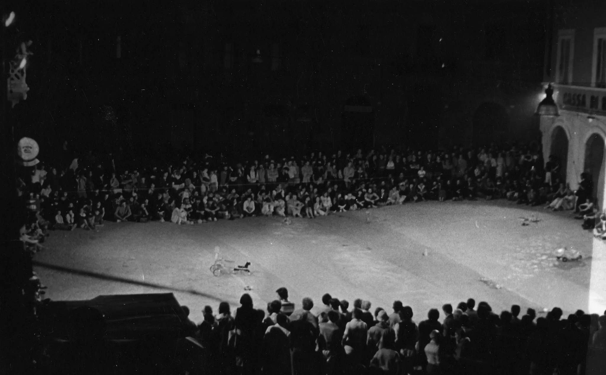 Przestrzeń miejska. Po dwóch stronach sceny stoją widzowie. Na scenie stoją ledwo widoczne przedmioty przypominające pojazdy mechaniczne.
