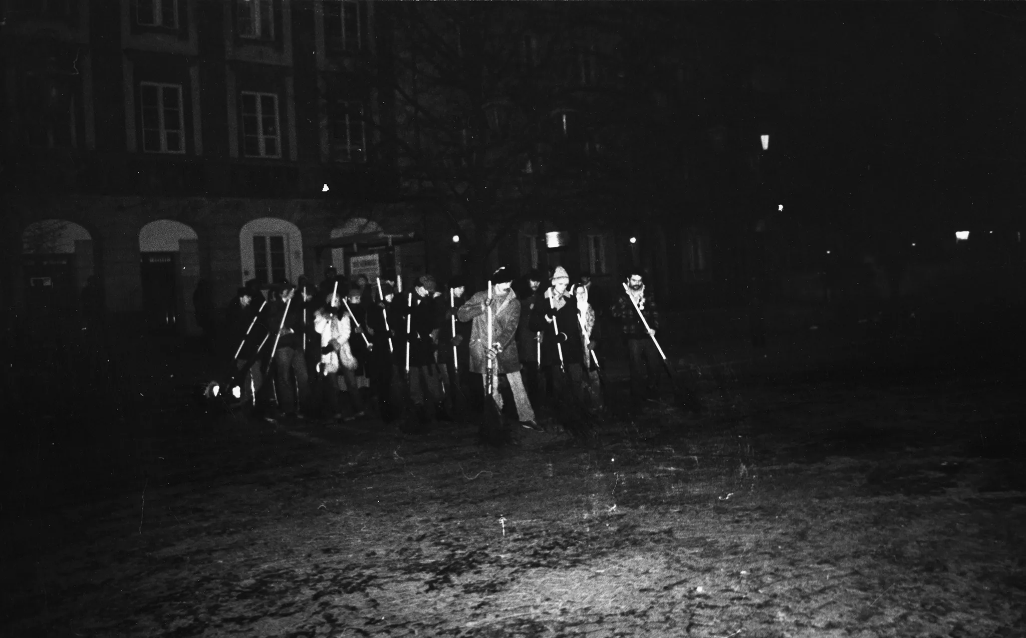 Niewyraźne zdjęcie. Przestrzeń miejska. Grupa aktorów i aktorek stoi na trawniku. Trzymają w rękach coś, co przypomina miotły. Za ich plecami stoi budynek.