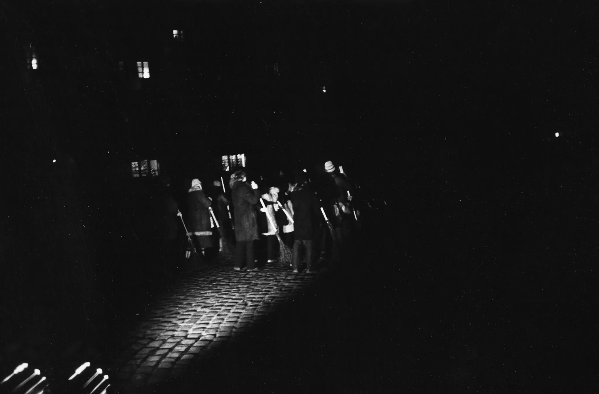 Niewyraźne zdjęcie. Przestrzeń miejska. Na brukowanej ulicy stoi grupa akorów i aktorek odwróconych do widza plecami. W rękach trzymają coś, co przypomina miotły. Oświetlają ich reflektory leżące na bruku.