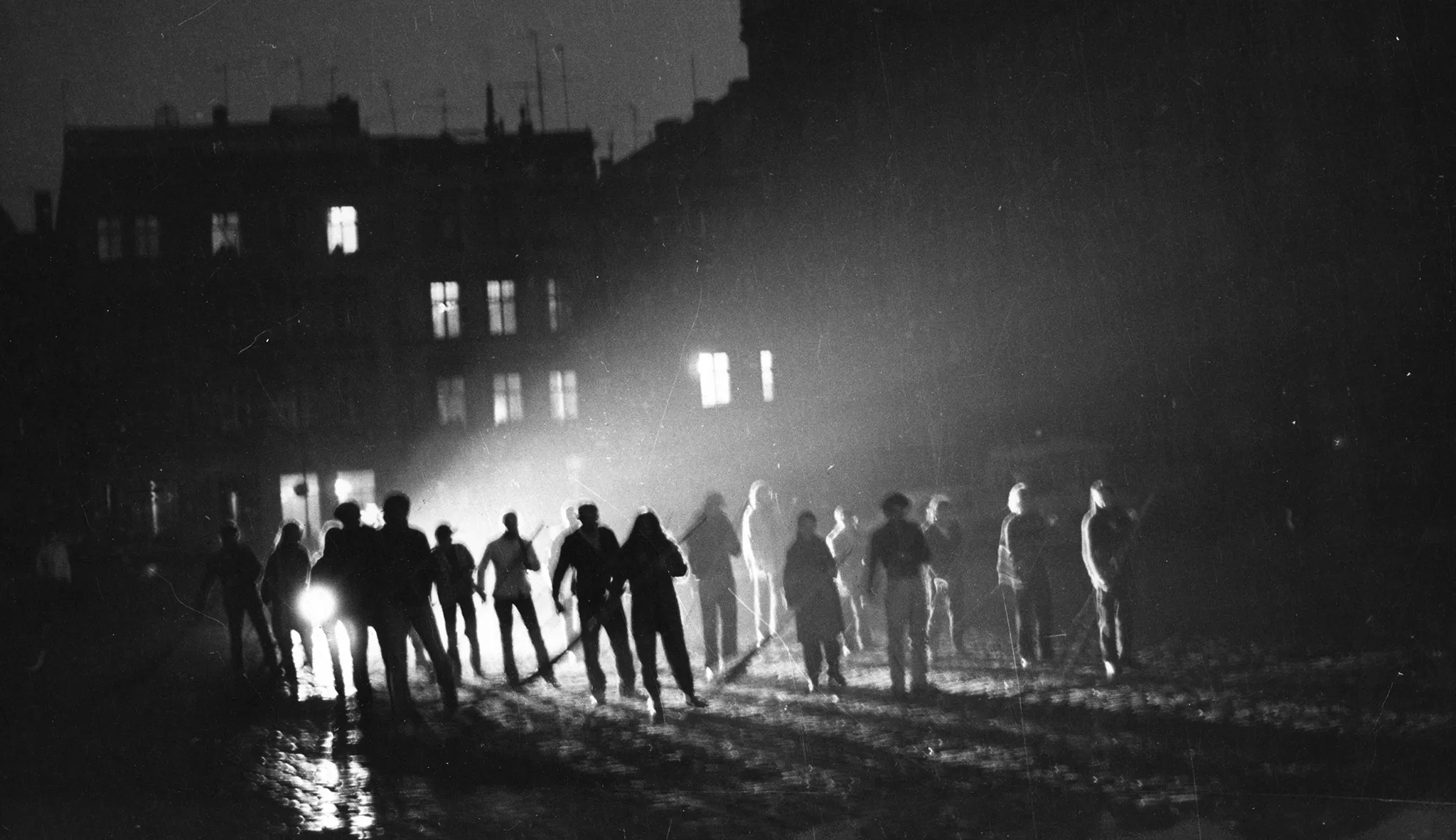Niewyraźne zdjęcie. Przestrzeń miejska. Między blokami stoi grupa aktorów i aktorek. W rękach trzymają coś, co przypomina miotły. Za ich plecami leżą reflektory, które oświetlają przestrzeń.