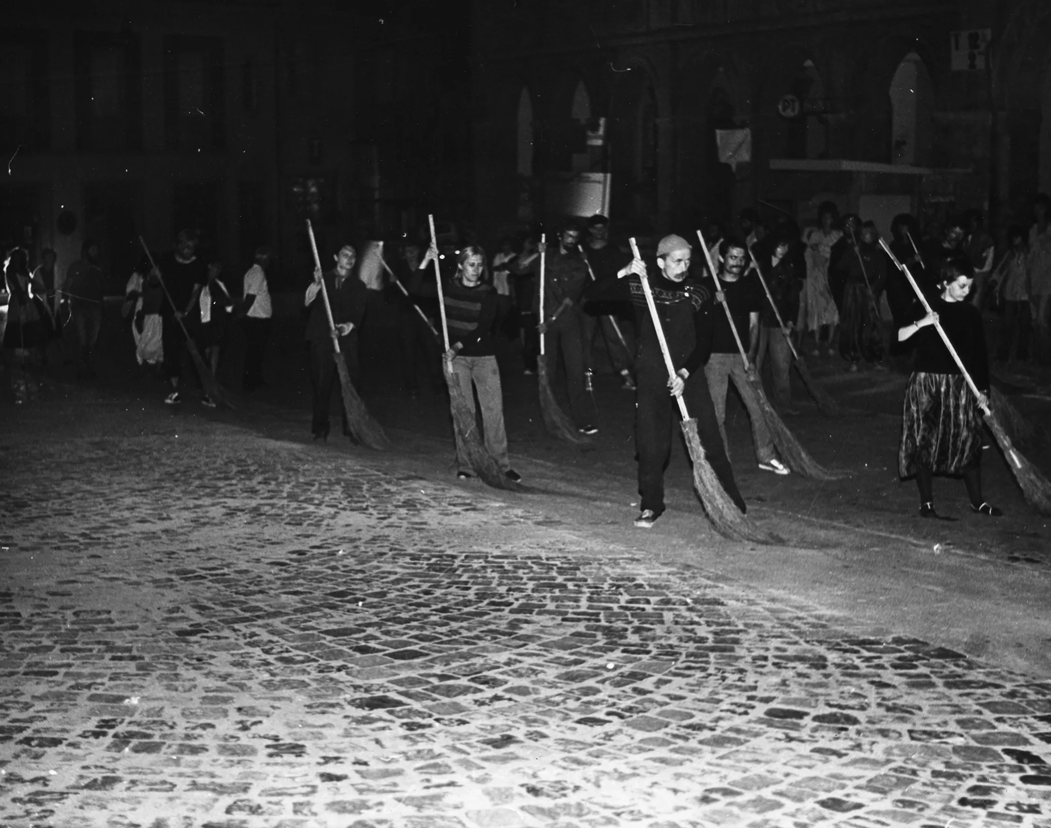 Przestrzeń miejska. Na brukowanej ulicy stoi grupa aktorów i aktorek. W rękach trzymają miotły, którymi zamiatają ulicę. Za ich plecami widać grupę widzów stojącą pod budynkiem.