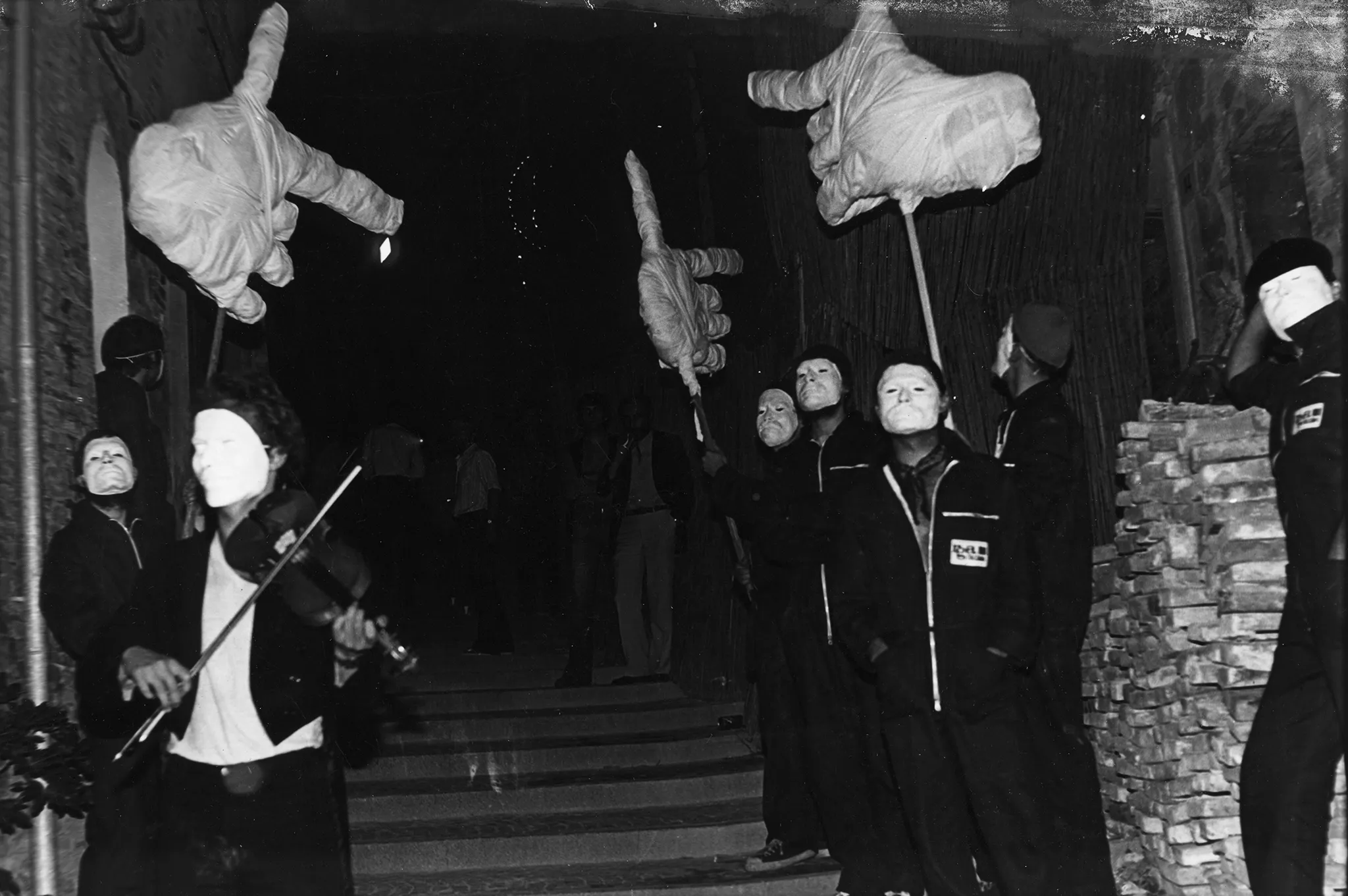 Przestrzeń miejska. Na schodach stoi grupa aktorów. Ubrani są na czarno, na twarzach mają białe maski teatralne. Niektórzy trzymają w rękach wielkie dłonie na kijach. Jeden z aktorów gra na skrzypcach.