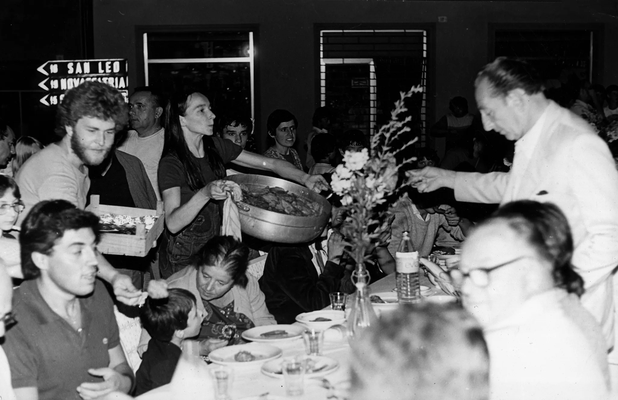 Wielki stół jadalny wystawiony na zewnątrz. Tłum aktorów i widzów. Trudno rozstrzygnąć, kto jest kim. W centrum stoi aktora z wielką miską pełną jedzenia. W tle widać budynek i kierunkowskazy drogowe.