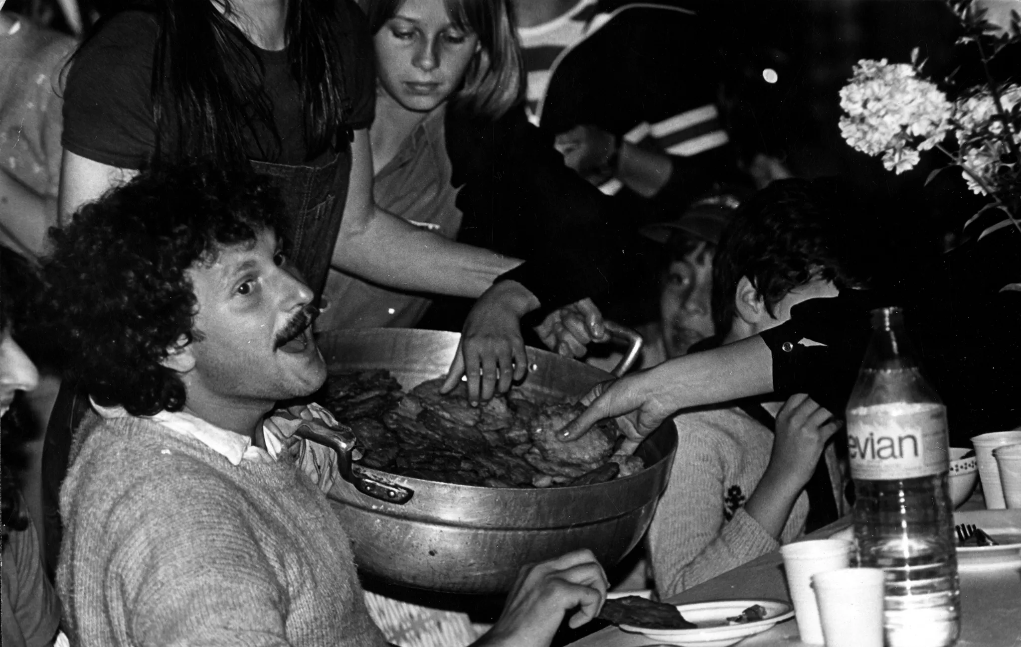 Przy stole siedzi aktor. W rękach trzyma miskę pełną jedzenia. Jedzenie wygląda jak placki. Wokół niego widać tłum ludzi.