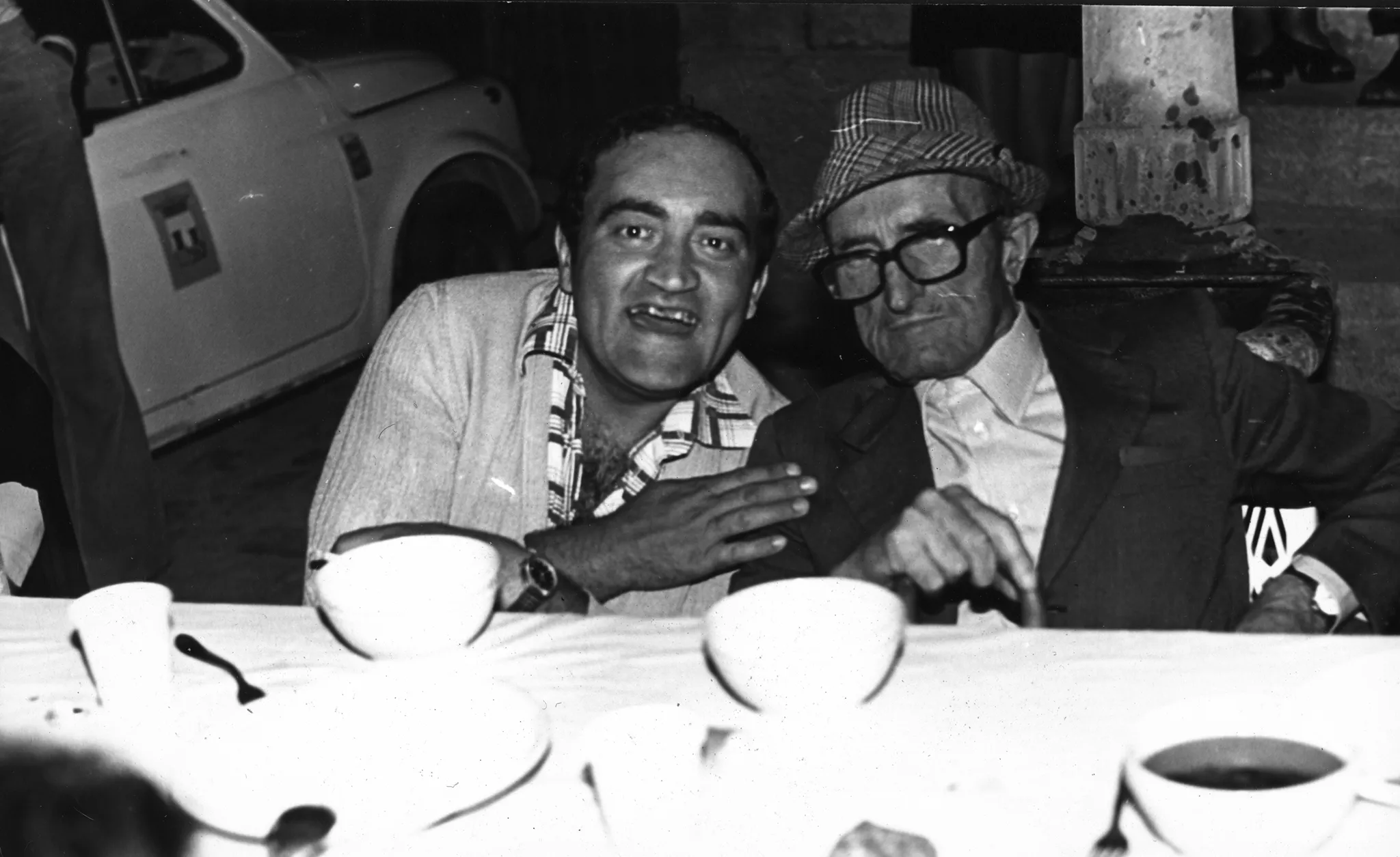 Przy stole siedzi dwóch mężczyzn patrzących w stronę widza. Pozują do zdjęcia. Mężczyzna z lewej strony ubrany jest w koszulę i szeroko się uśmiecha. Mężczyzna z prawej strony ubrany jest w garnitur, kapelusz i okulary optyczne. Jest poważny. Na stole stoją filiżanki z kawą.