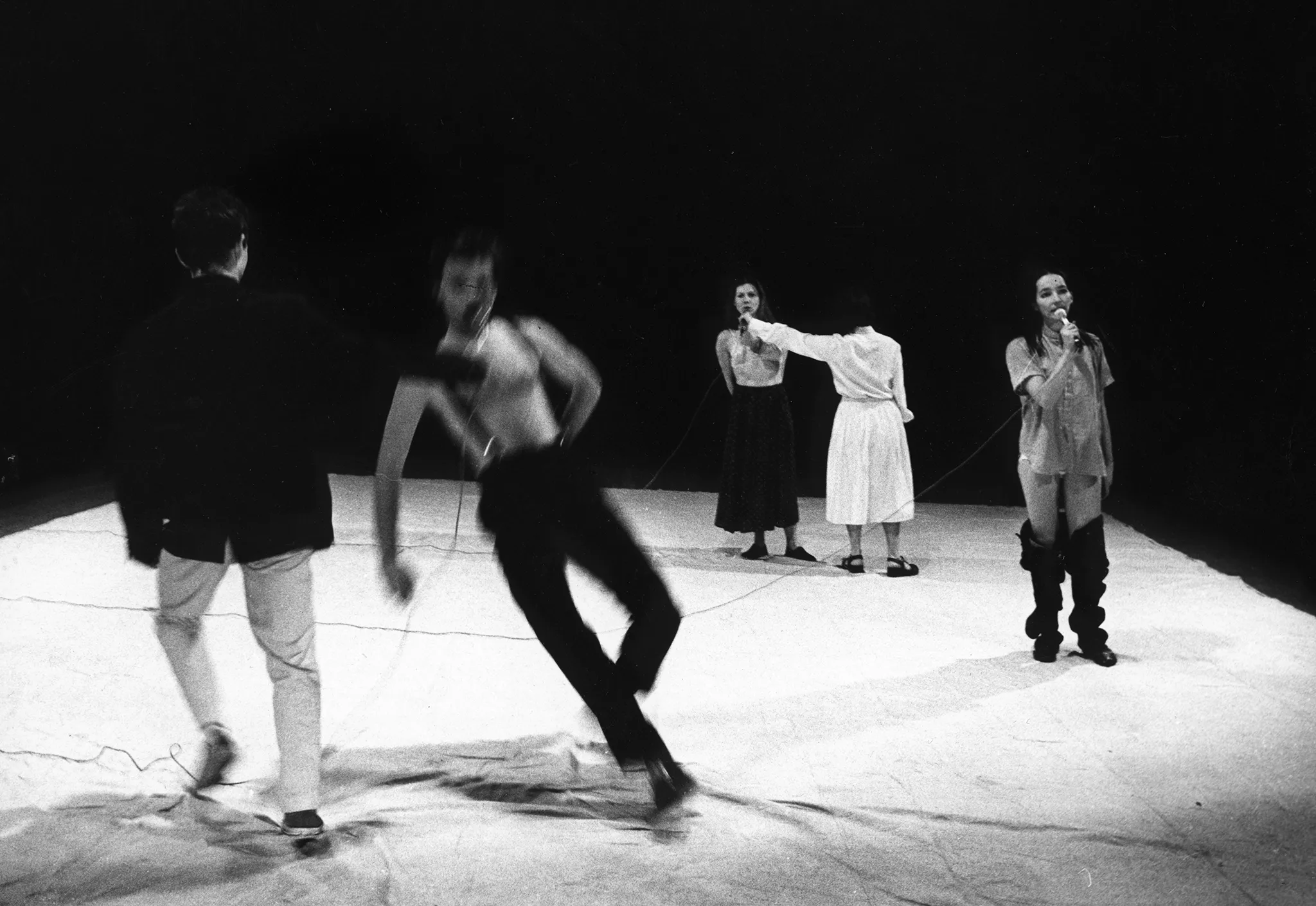 Na białej płachcie scenie stoi pięcioosobowa grupa aktorów i aktorek. Z przodu stoi zwrócony plecami do widza aktor trzymający mikrofon przy zwróconym twarzą w stronę widza aktorze, który się przewraca. Za nimi stoi zwrócona twarzą w stronę widza aktorka z mirkfonem. Ubrana jest w koszulę i wygląda, jakby spadły jej spodnie. Z tyłu stoi zwrócona plecami do widza aktorka trzymająca mikrofon przy aktorce zwróconej twarzą w stronę widza.