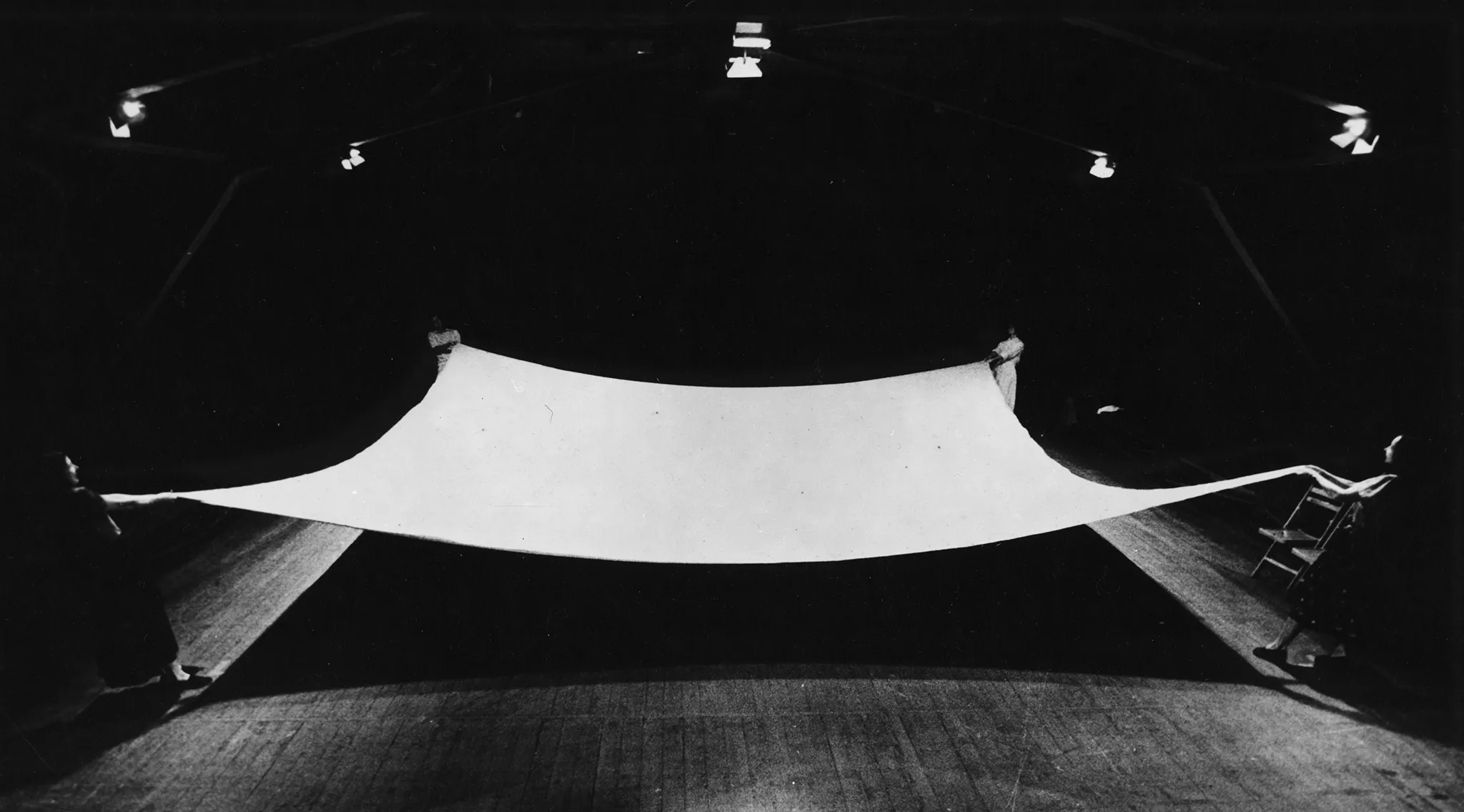 Na scenie stoi czteroosobowa grupa aktorów i aktorek. Każdy trzyma róg wielkiej białej kwadratowej płachty i unoszą ją nad scenę.