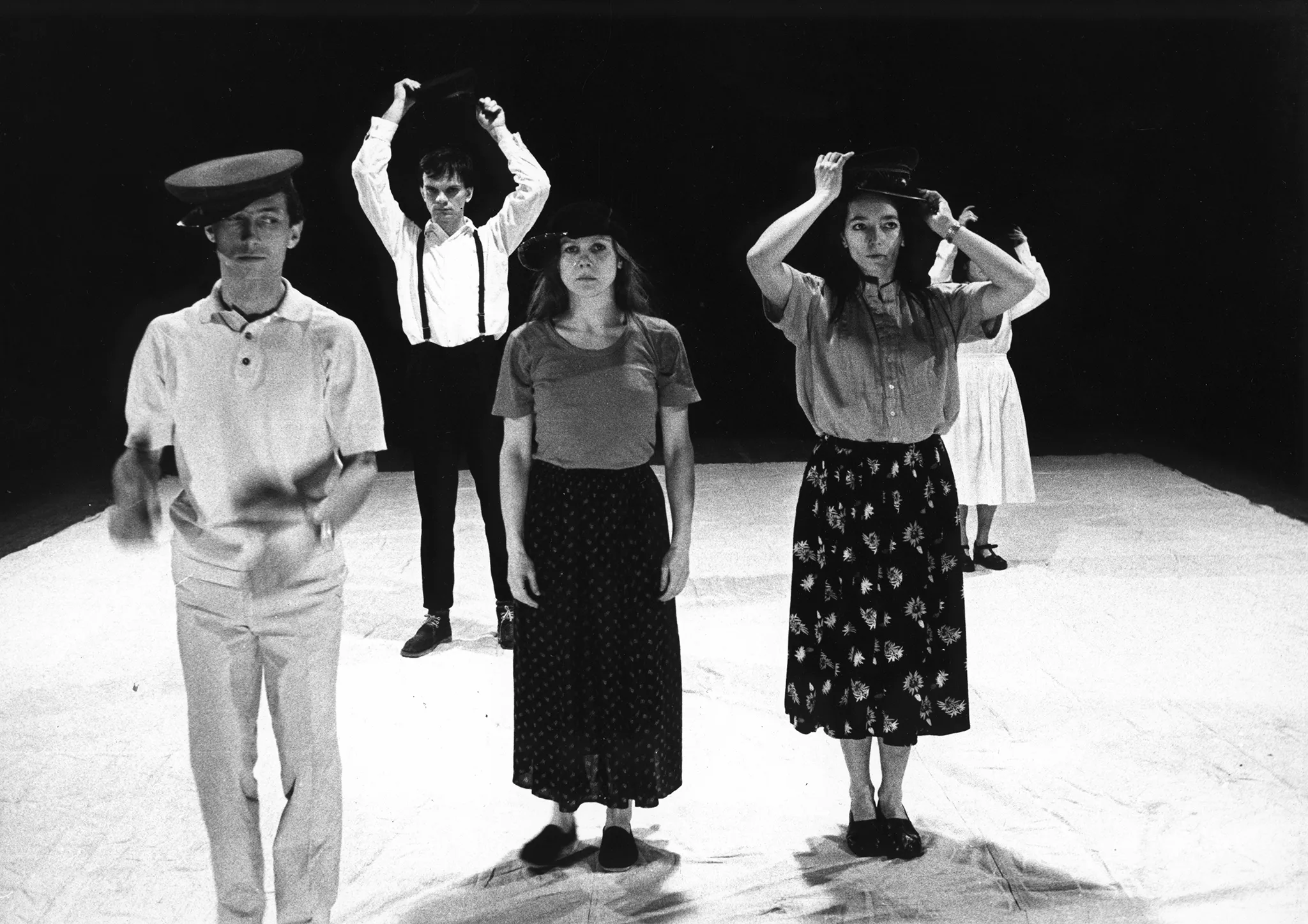 Na białej płachcie stoi pięcioosoba grupa aktorów i aktorek zwróconcych twarzą w stronę widza. Wyglądają, jakby byli ubrani prywatne. Wzrok przykuwają ich duże nakrycia głowy.