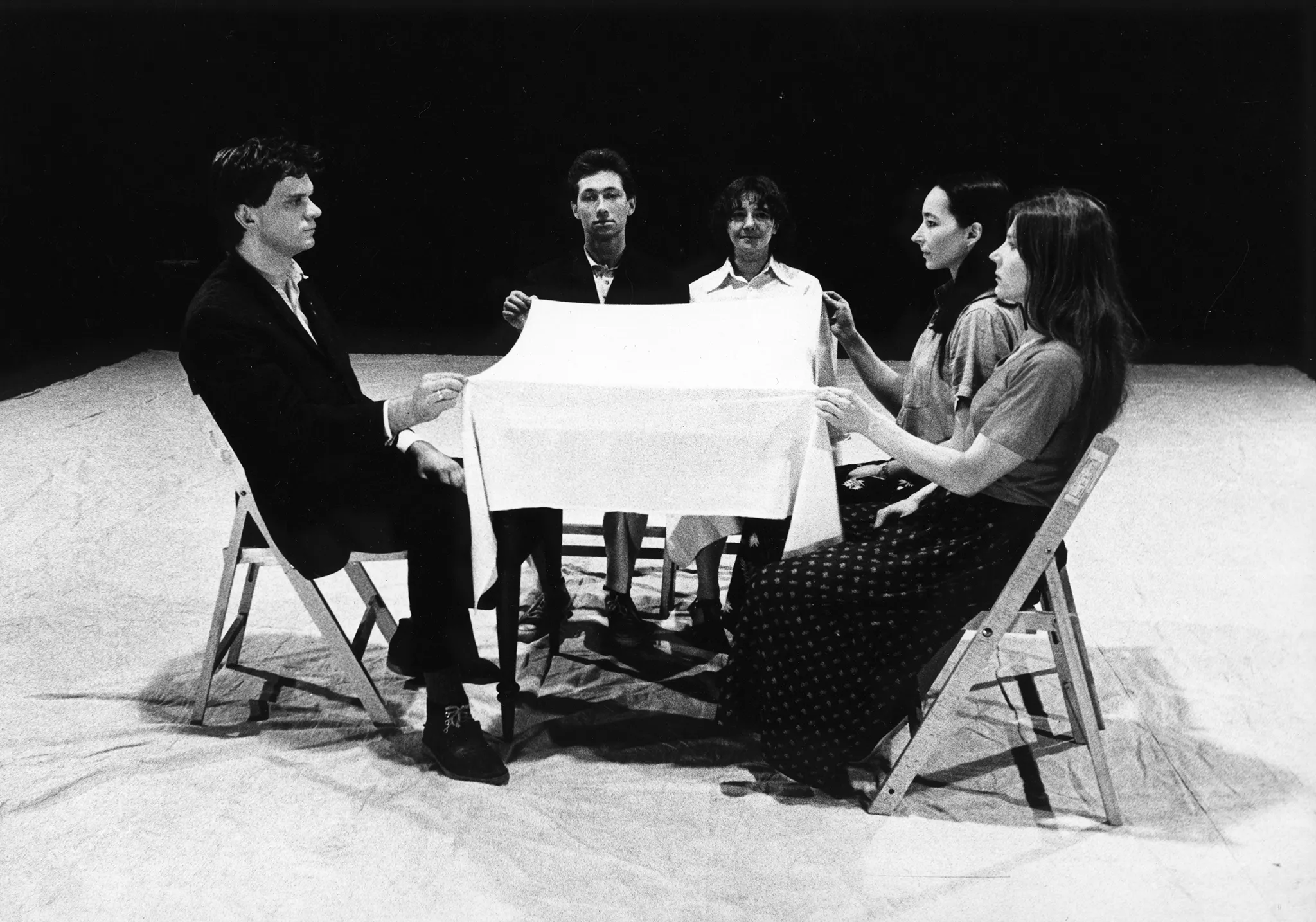 Przy stoliku siedzi pięcioosobowa grupa aktorów i aktorek. Trzymają w rękach biały obrus i unoszą go nad stołem.