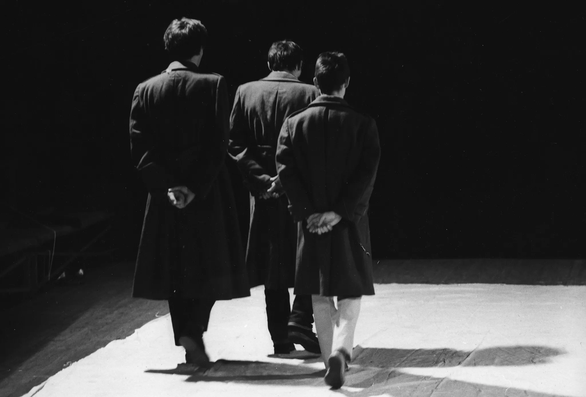 Trzech aktorów zwróconych plecami w stronę widza. Wszyscy ubrani są w długie ciemne płaszcze. Powoli idą przed siebie.